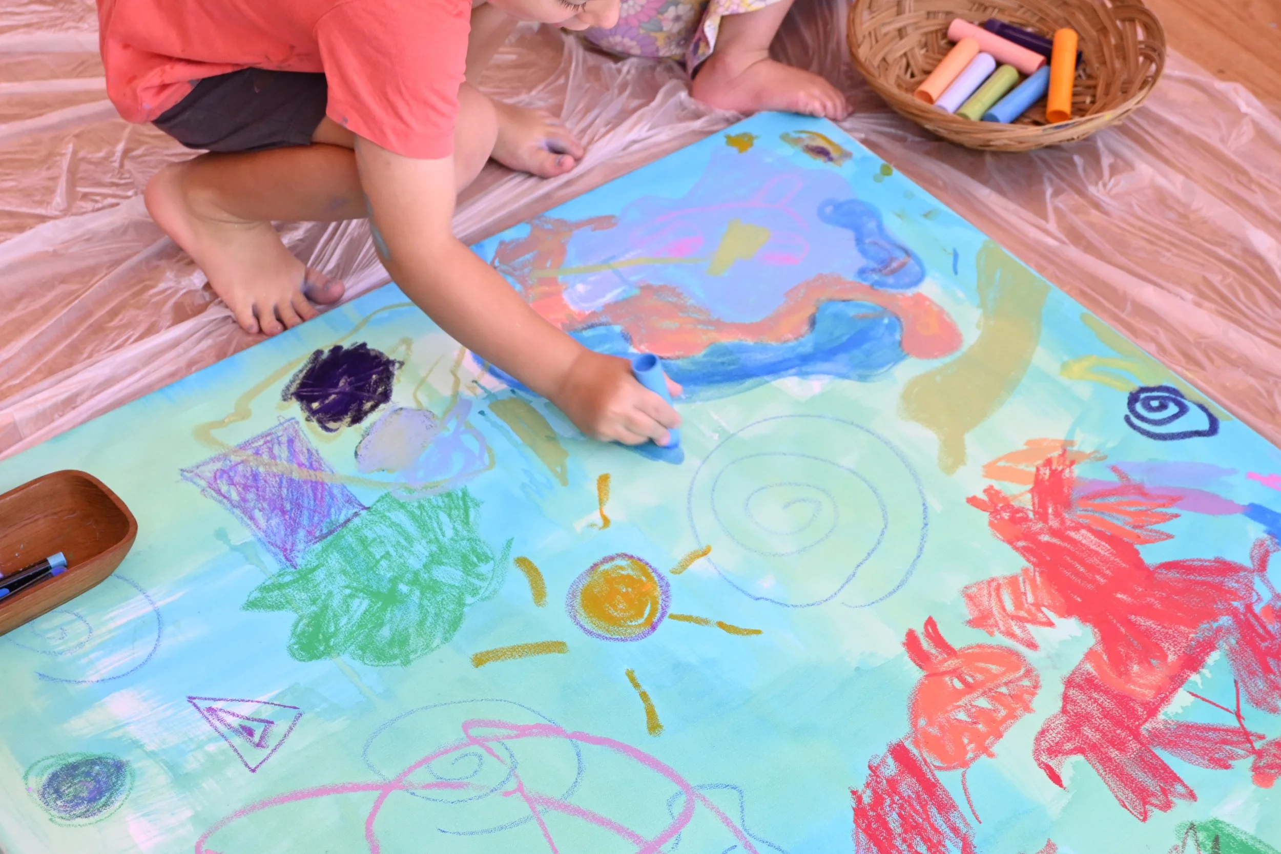 A child drawing colorful shapes and scribbles on a large sheet of paper with chalk inside a room. The paper features a drawing of the sun, a tree, and abstract designs. The child is kneeling on a plastic sheet, with a basket of chalk in front.