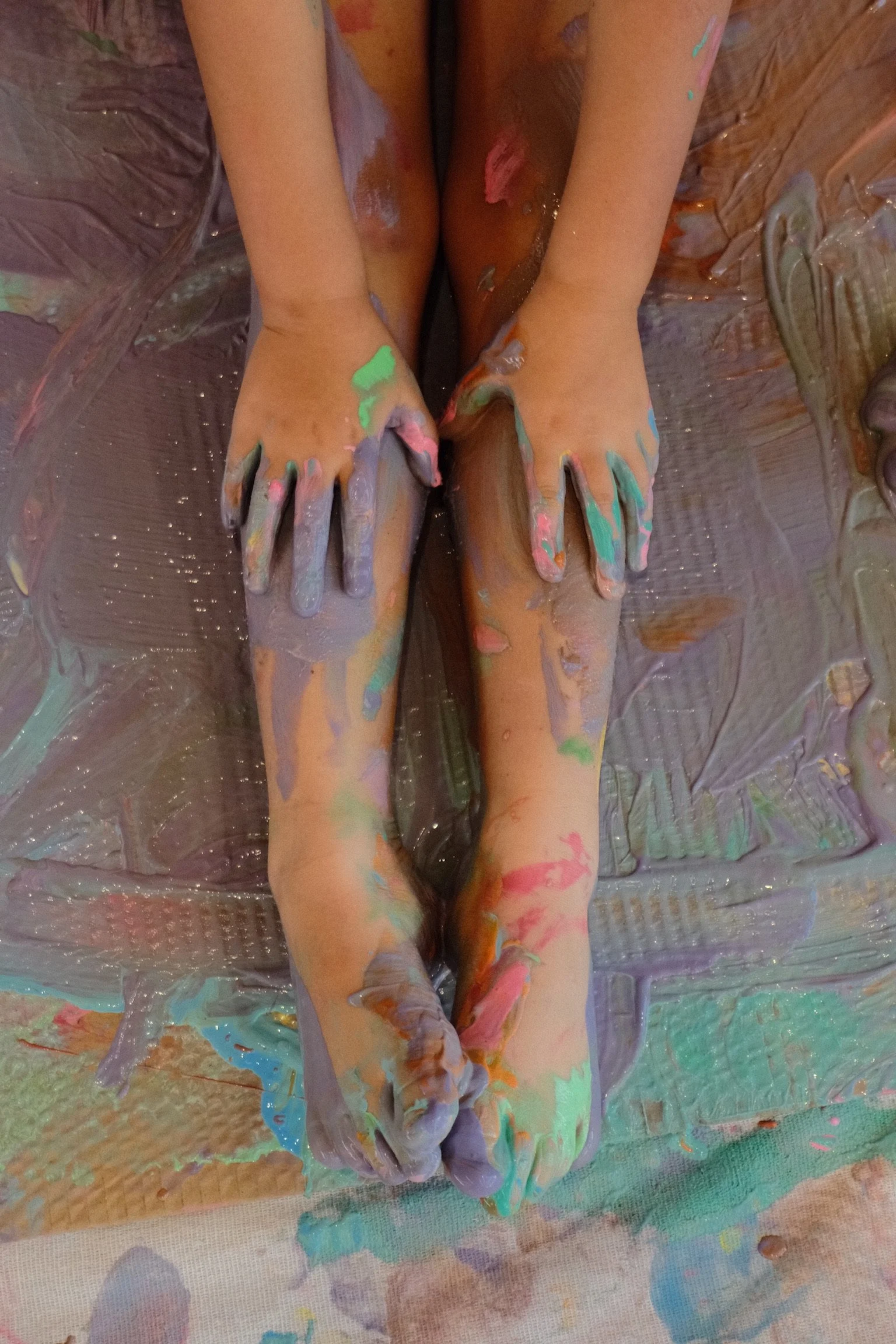 Child's legs and hands covered in multicolored paint, lying on a paint-splattered surface.