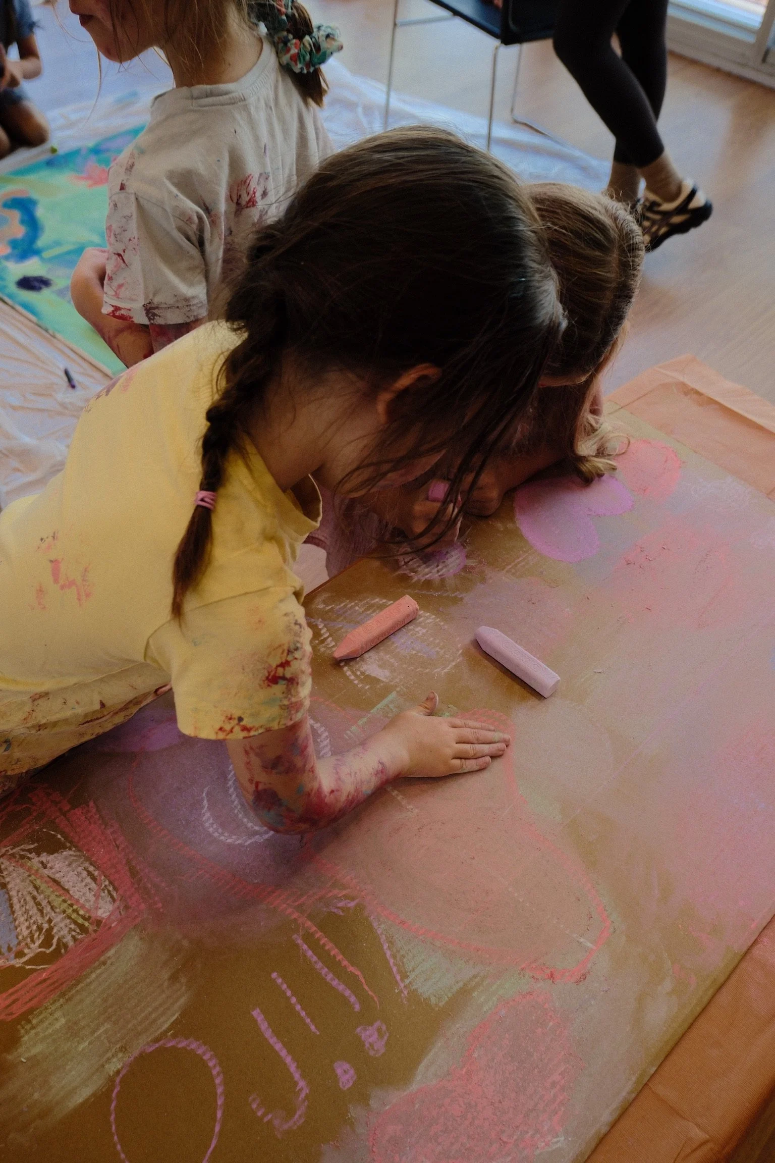 Children drawing on large paper with chalk or pastel, creating colorful heart and flower shapes.