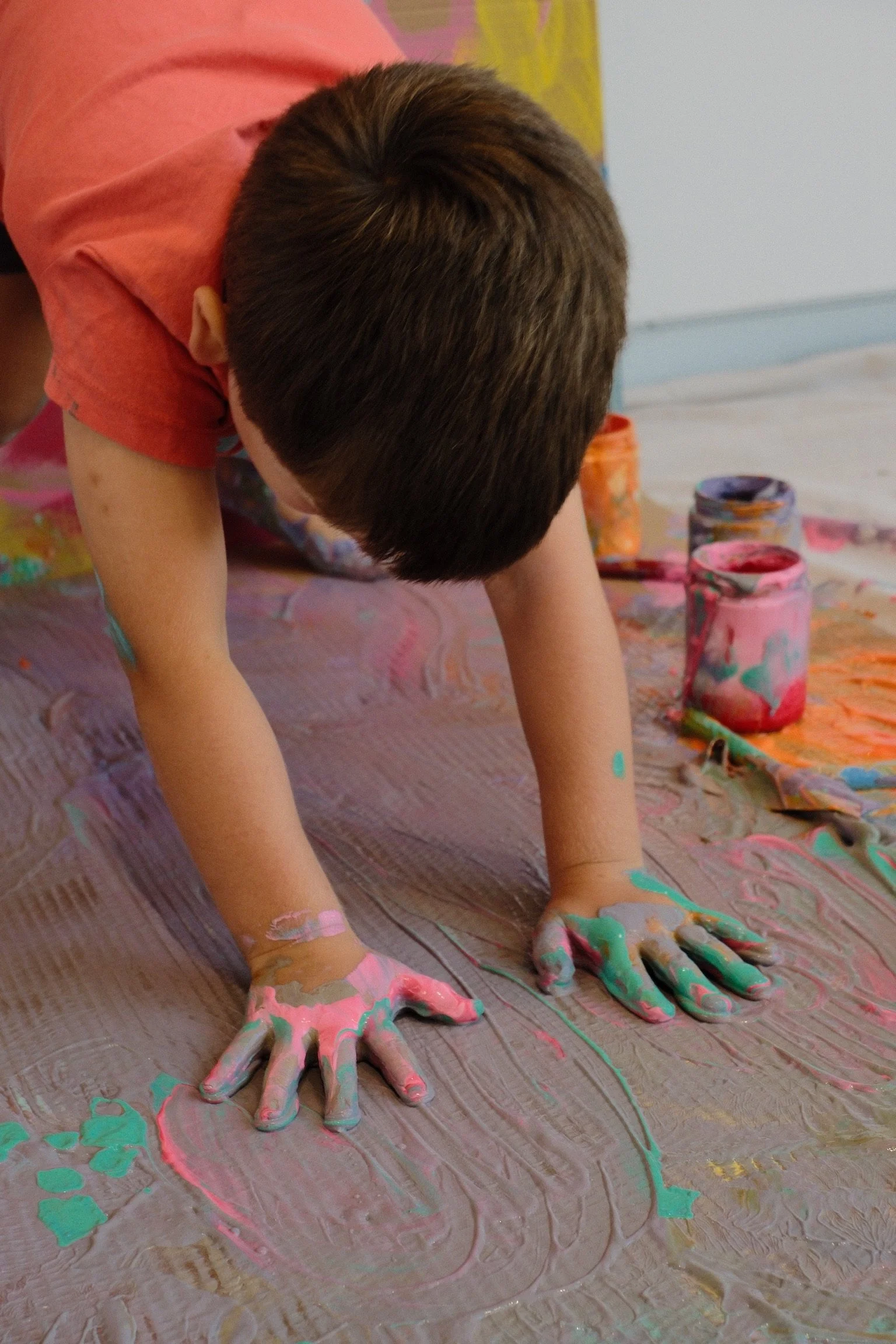A young boy is crawling on the floor, covered in colorful paint, with open jars of paint and a paintbrush nearby.