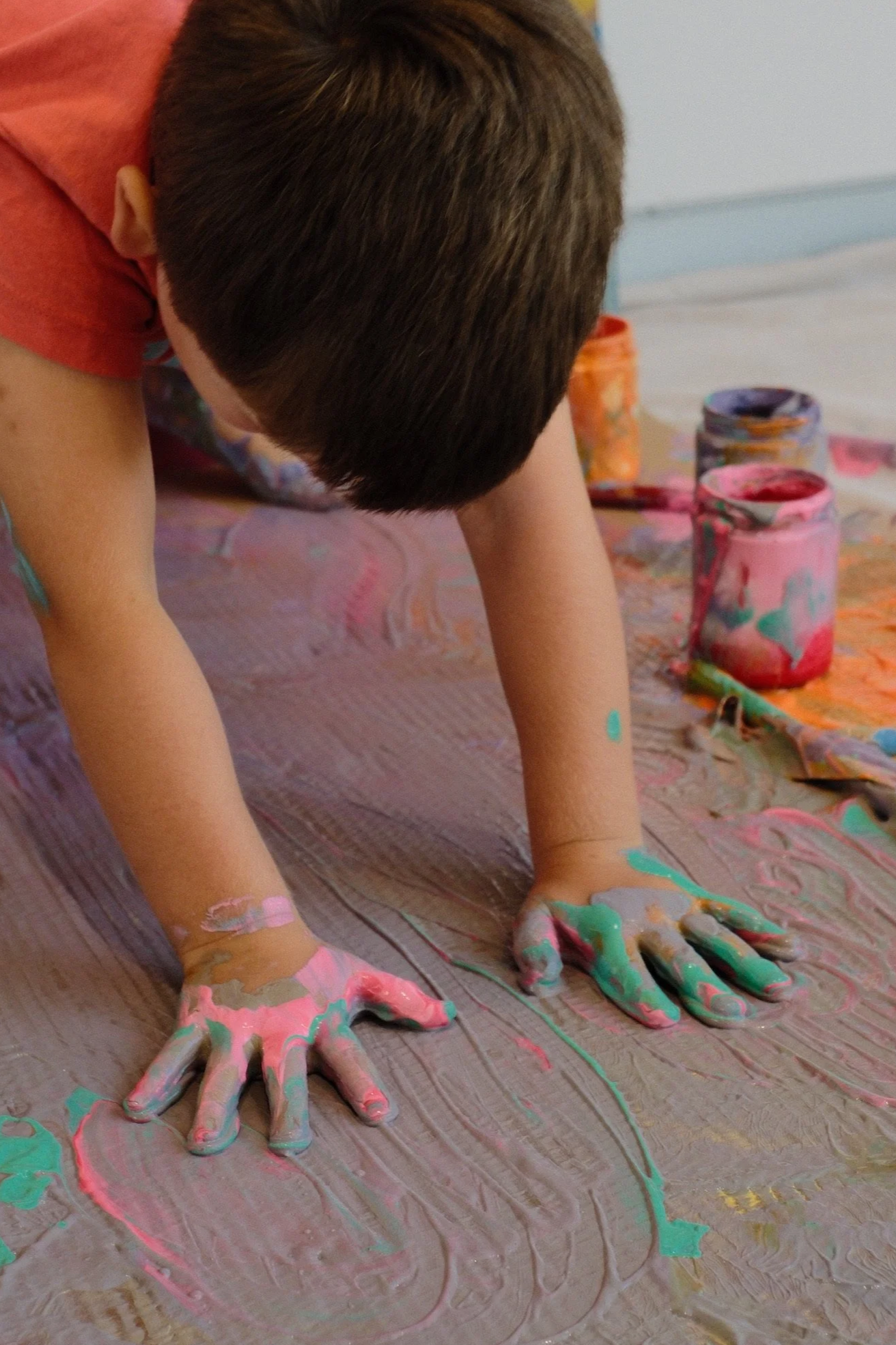 A child with dark hair and wearing a red shirt is on the floor, using both hands to mix multicolored paint on a large surface covered with brown paper. Several open paint jars with pink, purple, and green paint are in the background.