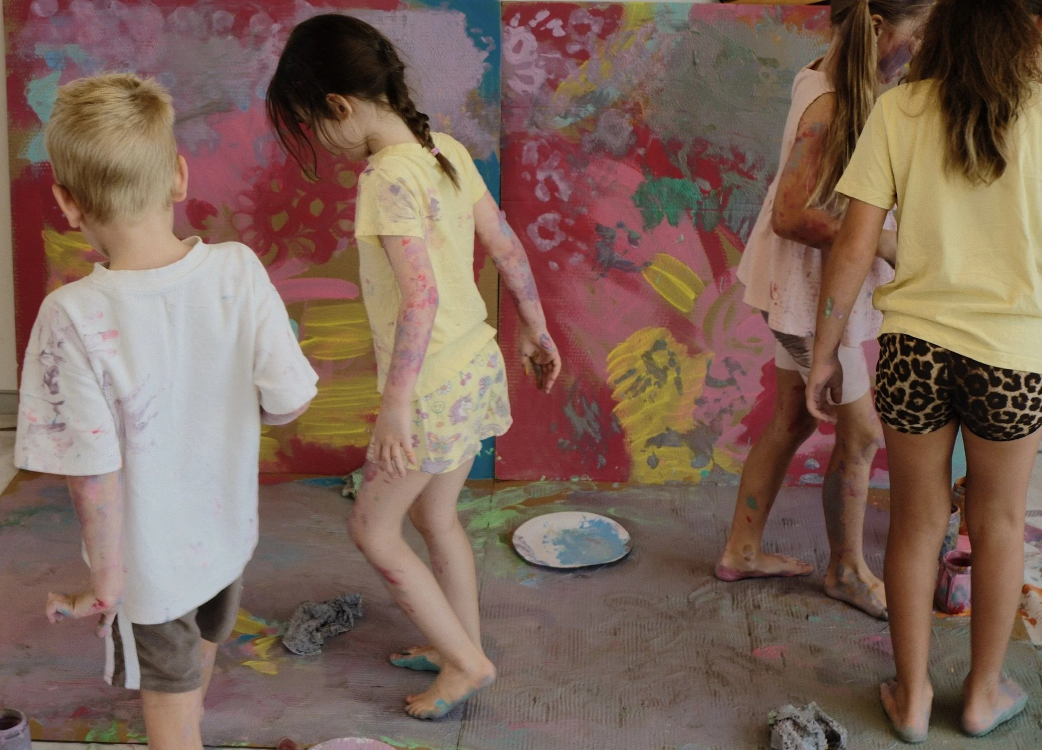 Children creating art on a large wall with colorful paint, some with paint-covered legs and clothes, in a room with a painted background.