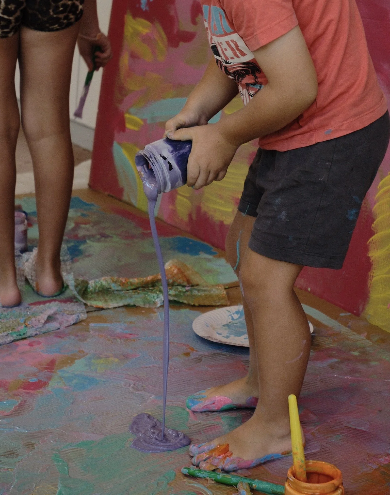 Child pouring purple paint onto colorful floor, surrounded by children in a messy, colorful art space.