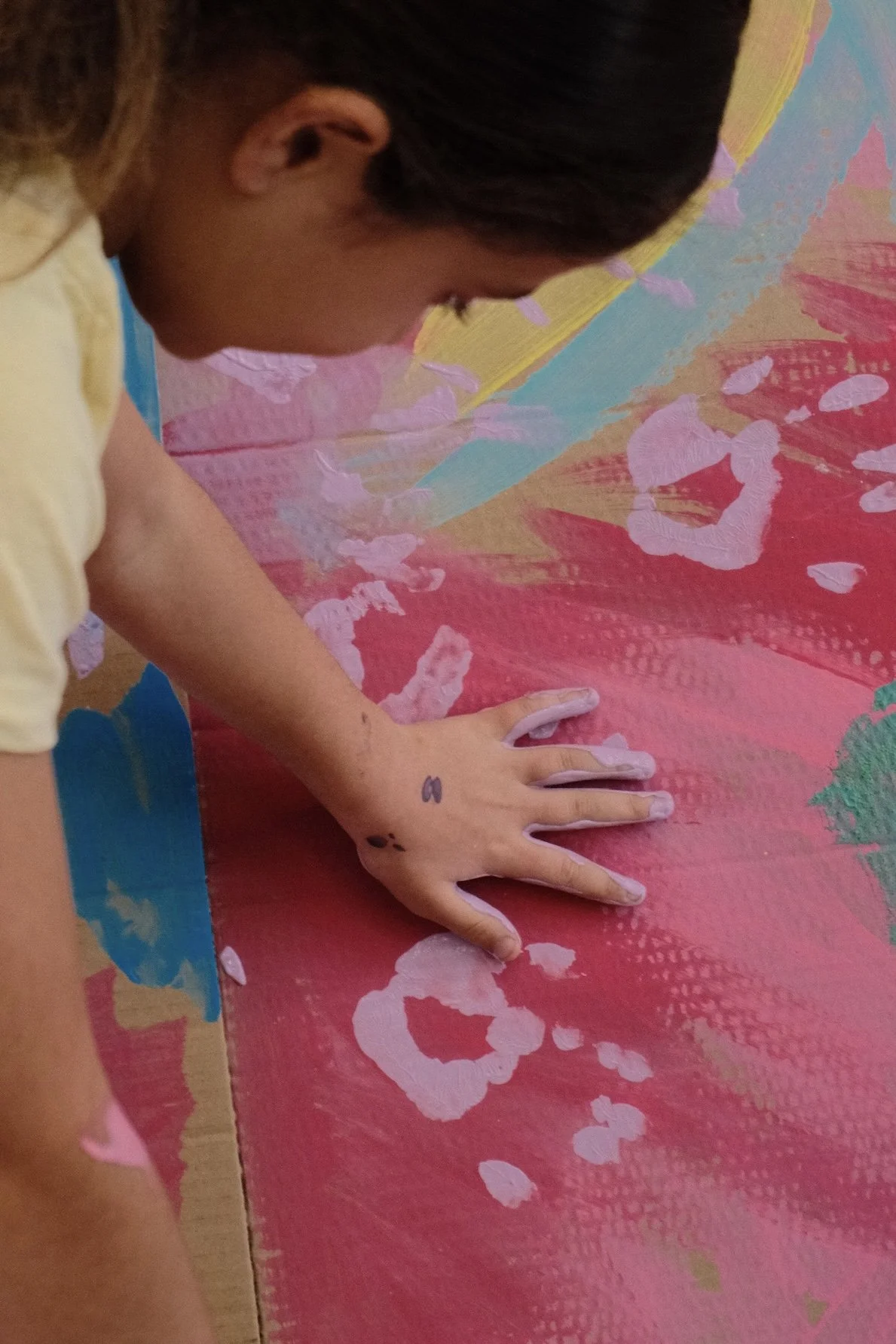 A person with dark hair, wearing a light-colored shirt, is creating an abstract painting on a colorful canvas using white paint. The canvas has pink, blue, green, and yellow sections.
