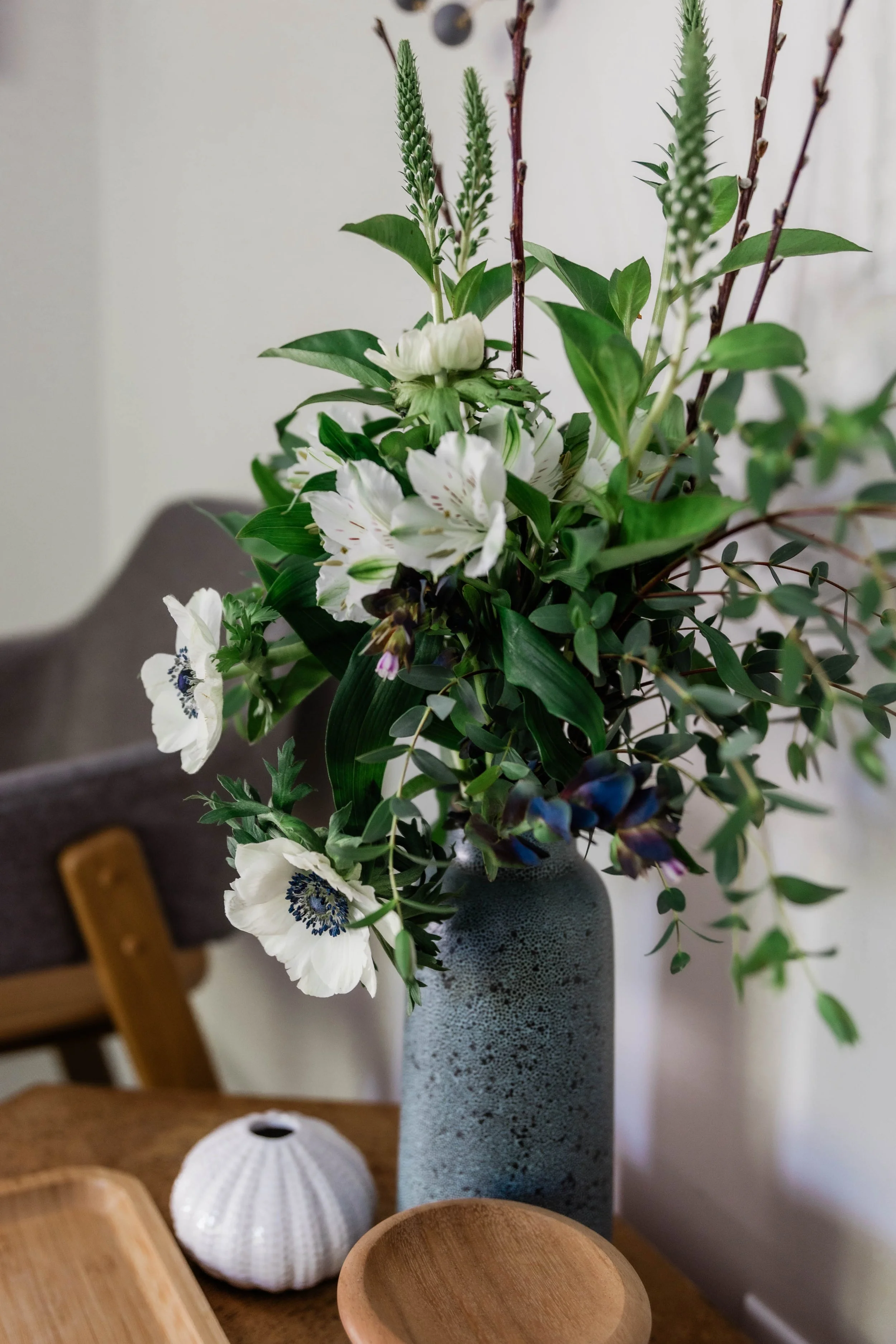 A bouquet of flowers in a textured gray vase on a table, with wooden bowls and decorative objects nearby.