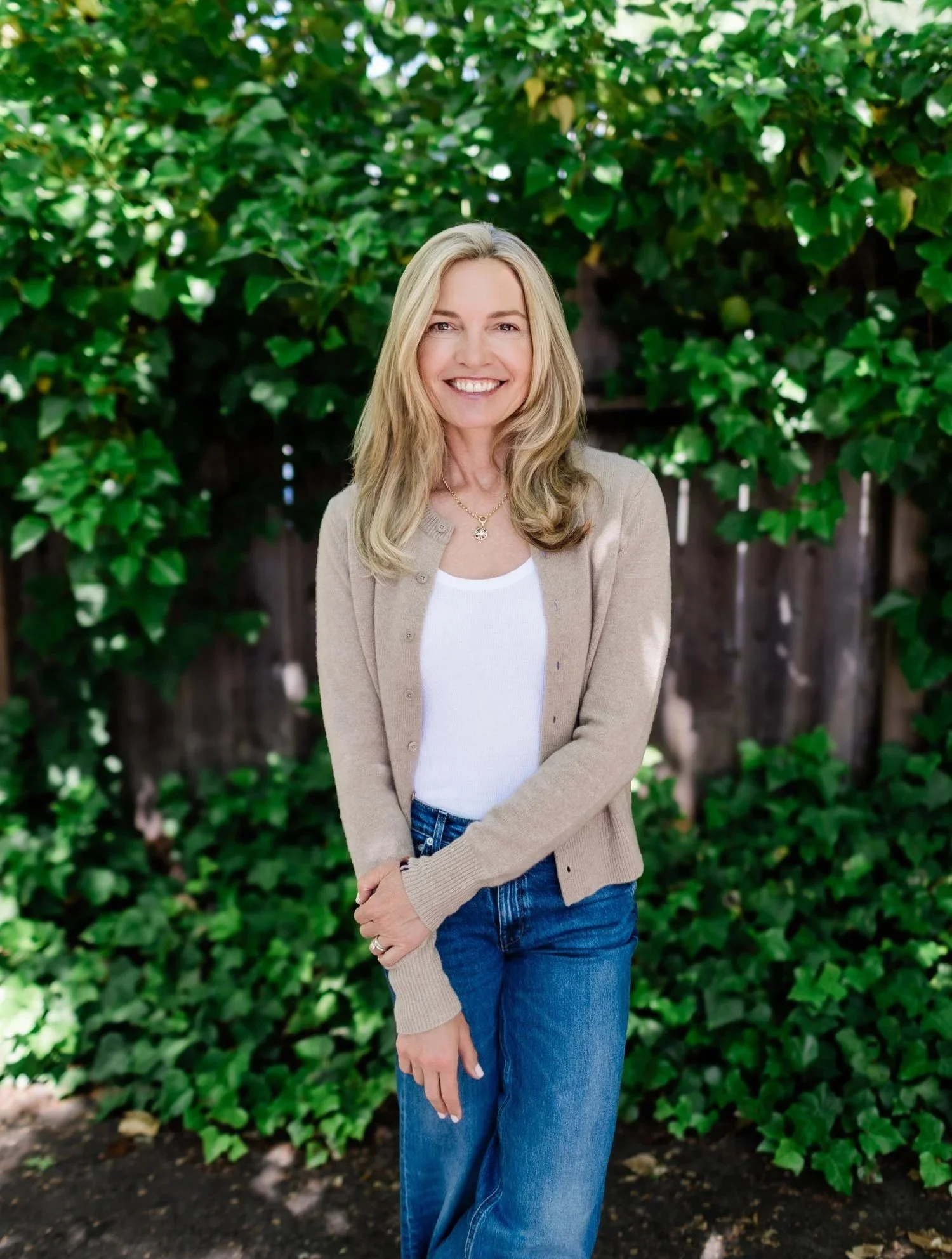 Smiling woman with blonde hair in a beige cardigan and jeans standing in front of a green leafy background.