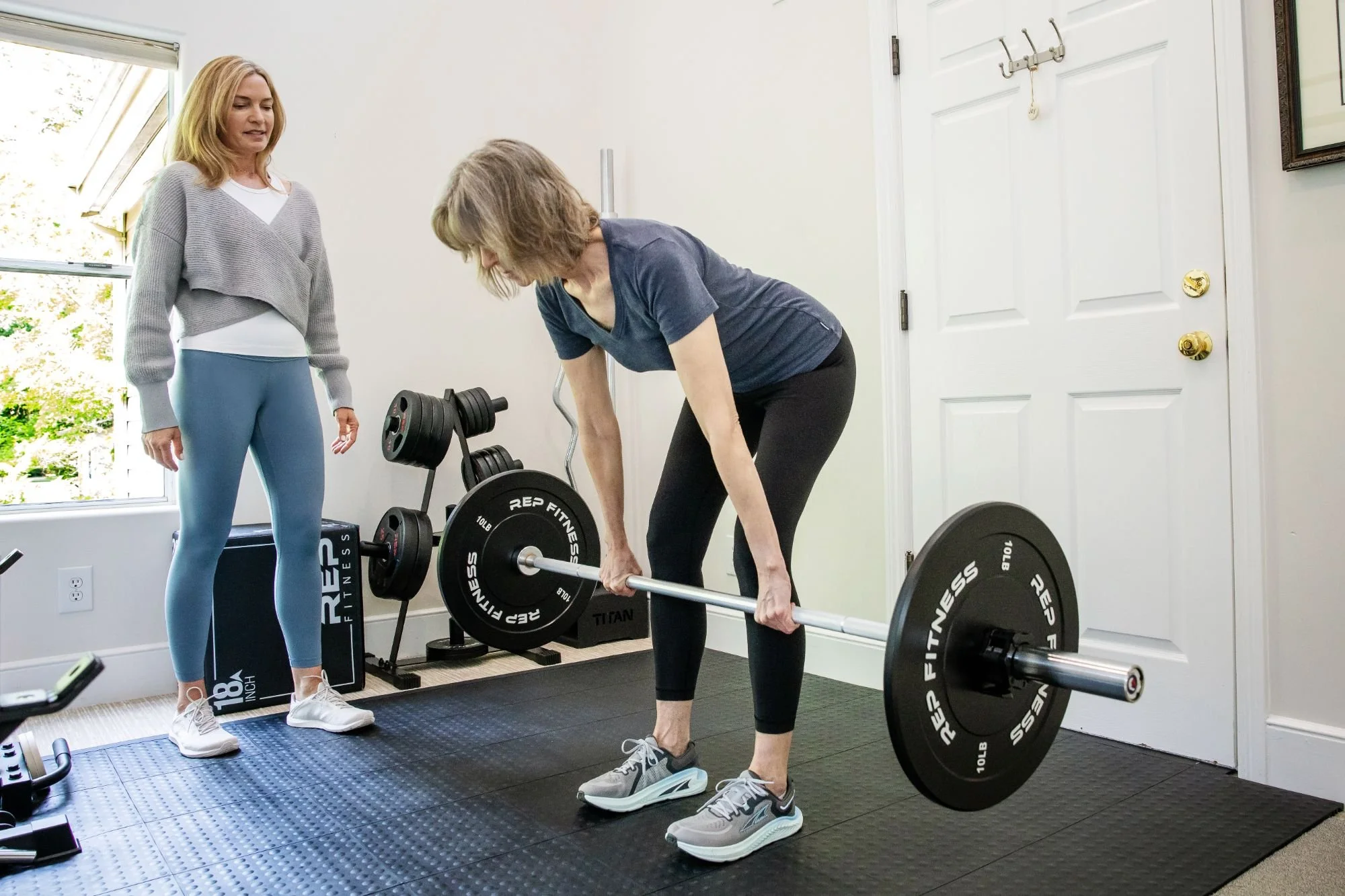 A woman in athletic shoes lifts a barbell in a home gym, assisted by another woman in workout attire who is observing. The space has a window, a coat hook, weights, and fitness equipment.