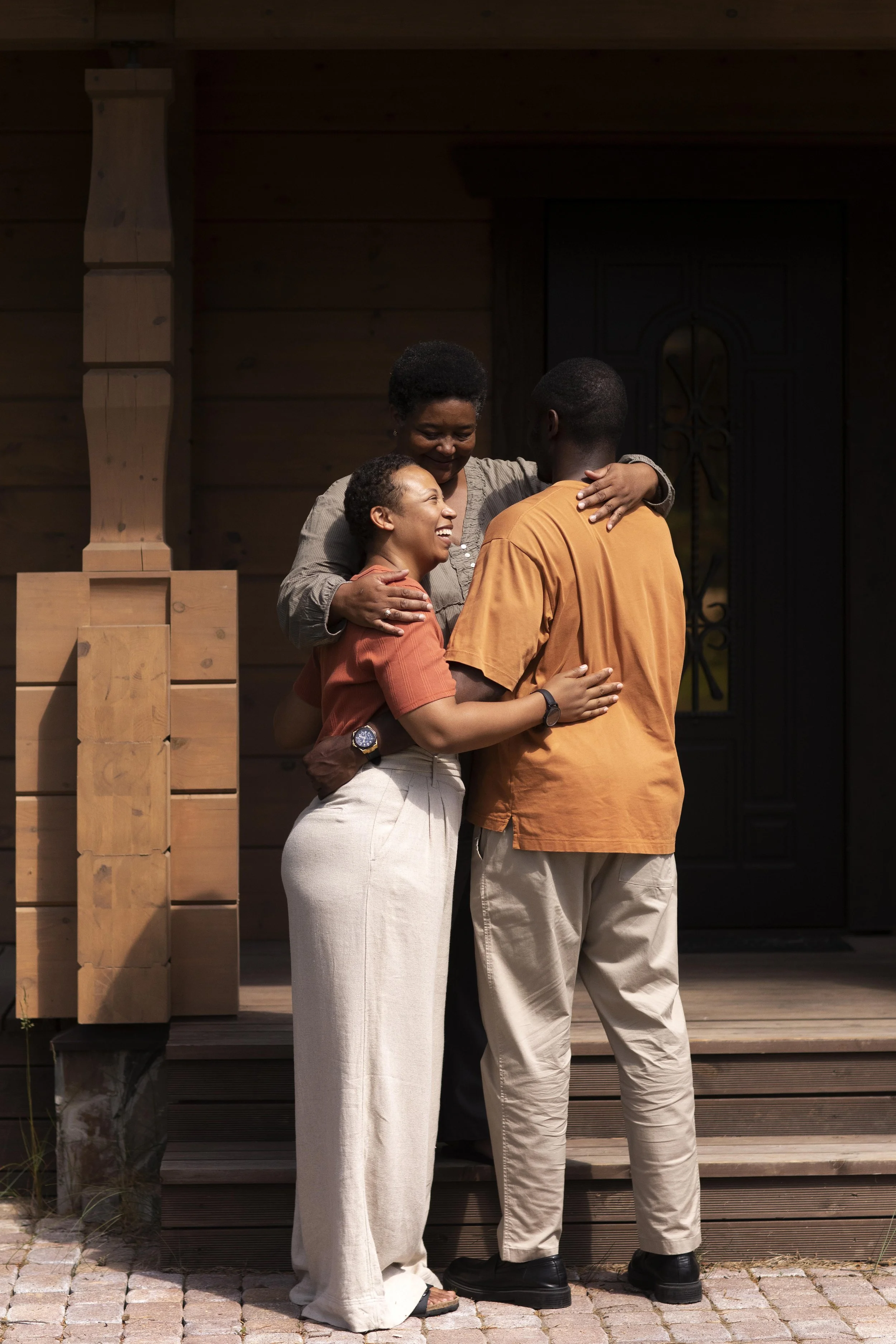 Three people hugging and smiling outside a wooden house, standing on wooden steps.