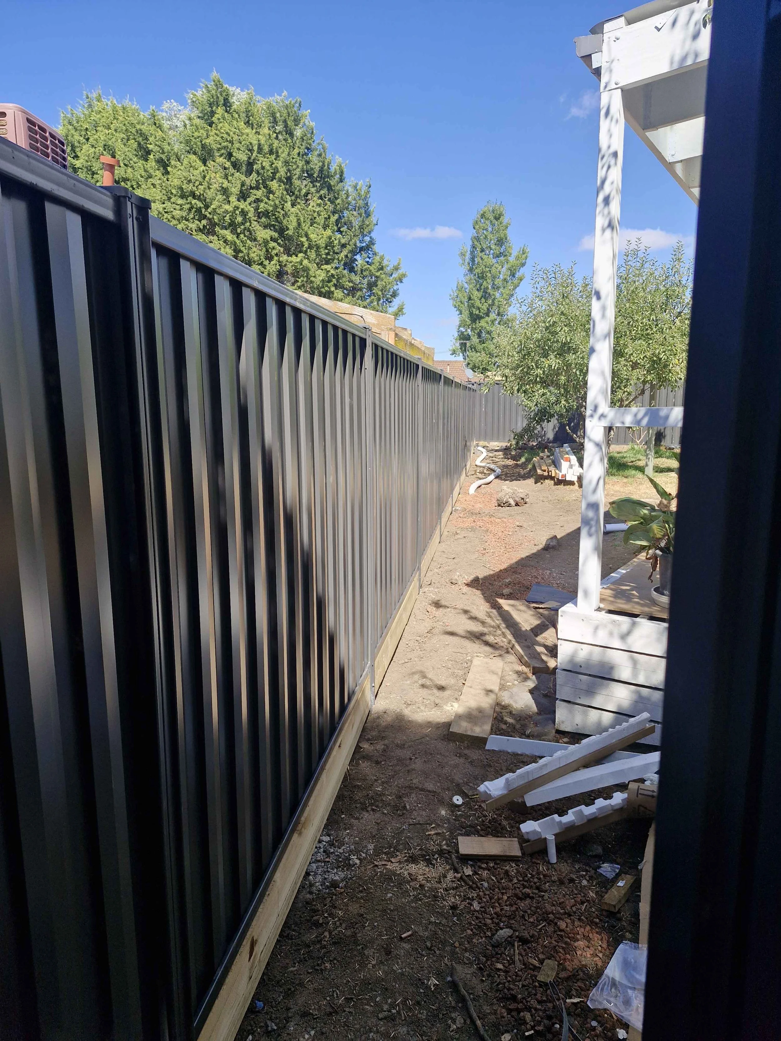 A backyard with a black metal fence, some construction materials, and a white wooden structure with a potted plant. Trees and blue sky with some clouds in the background.
