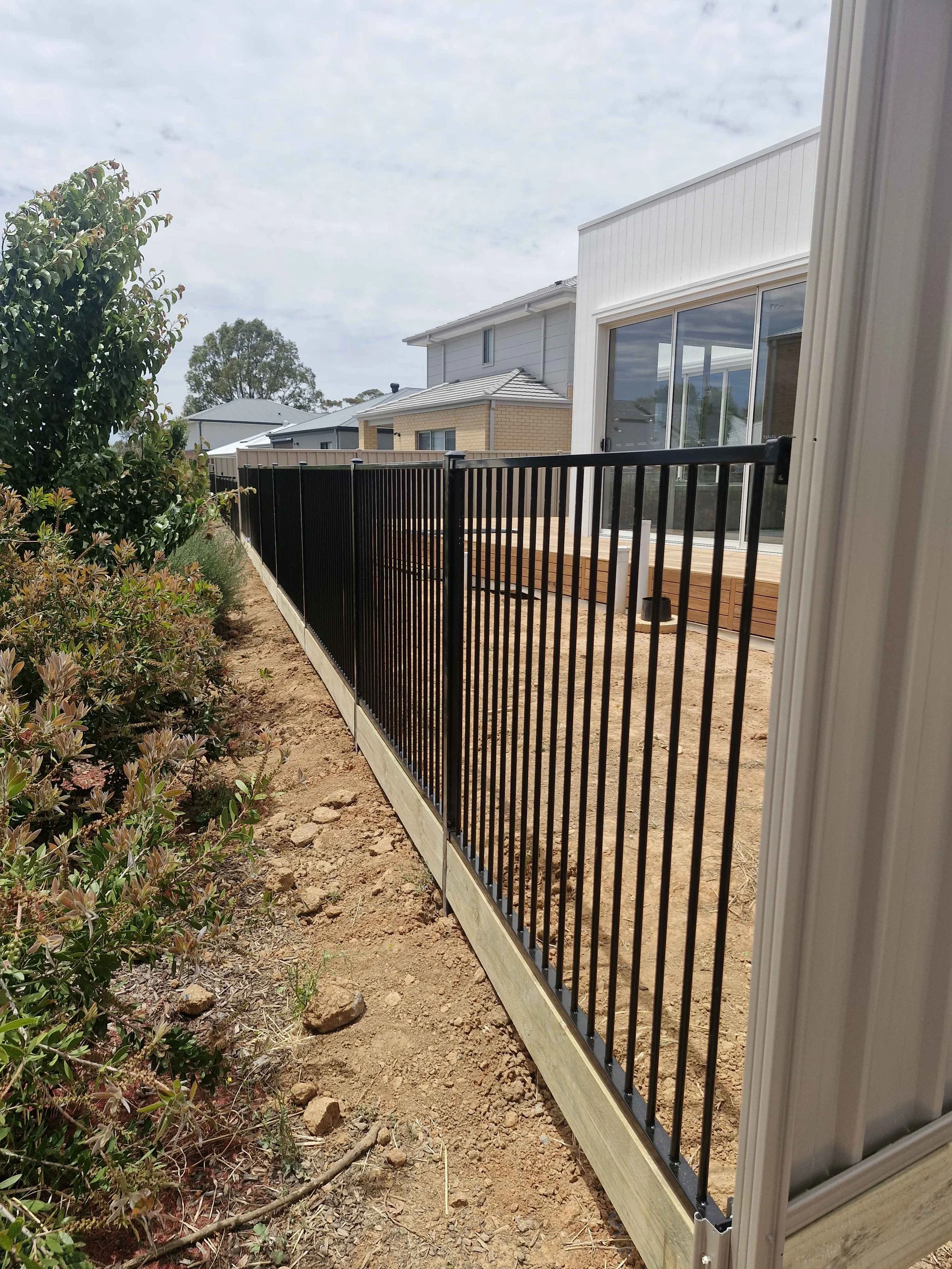 Construction of a black metal fence along a yard with soil and a few plants, in front of a house with large windows and neighboring houses in the background.