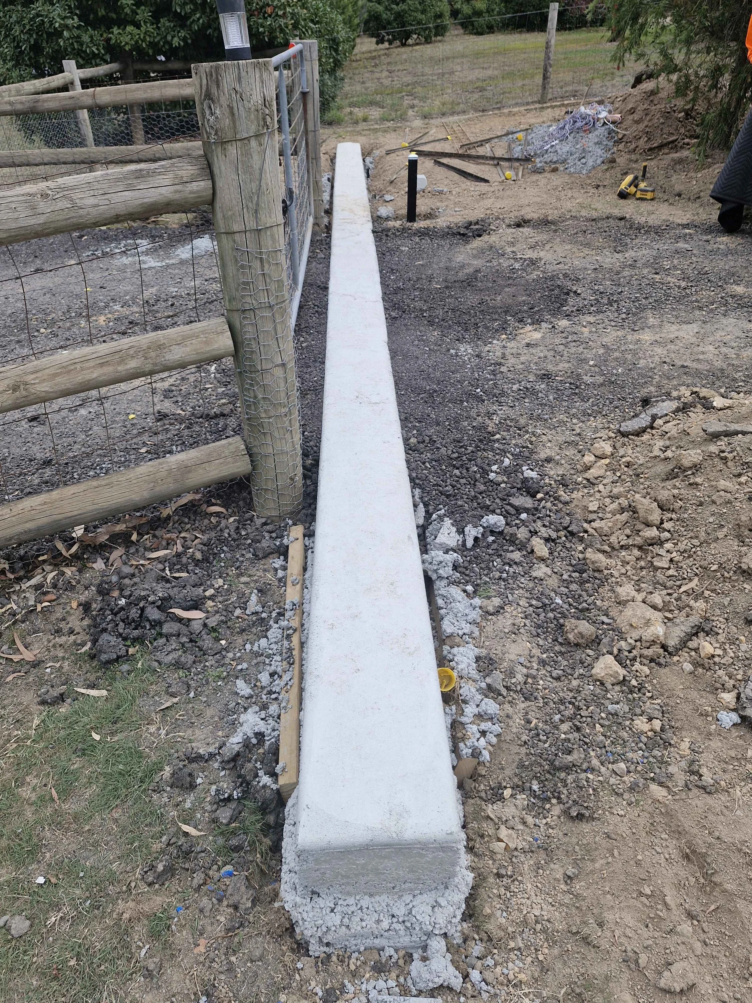 Long concrete curb being installed on a construction site beside a wooden fence, with tools and construction materials nearby.
