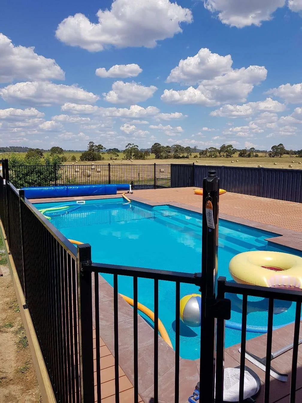 Small rectangular swimming pool with a pool float and water toys, surrounded by a black metal fence, with a field and trees under a partly cloudy sky in the background.