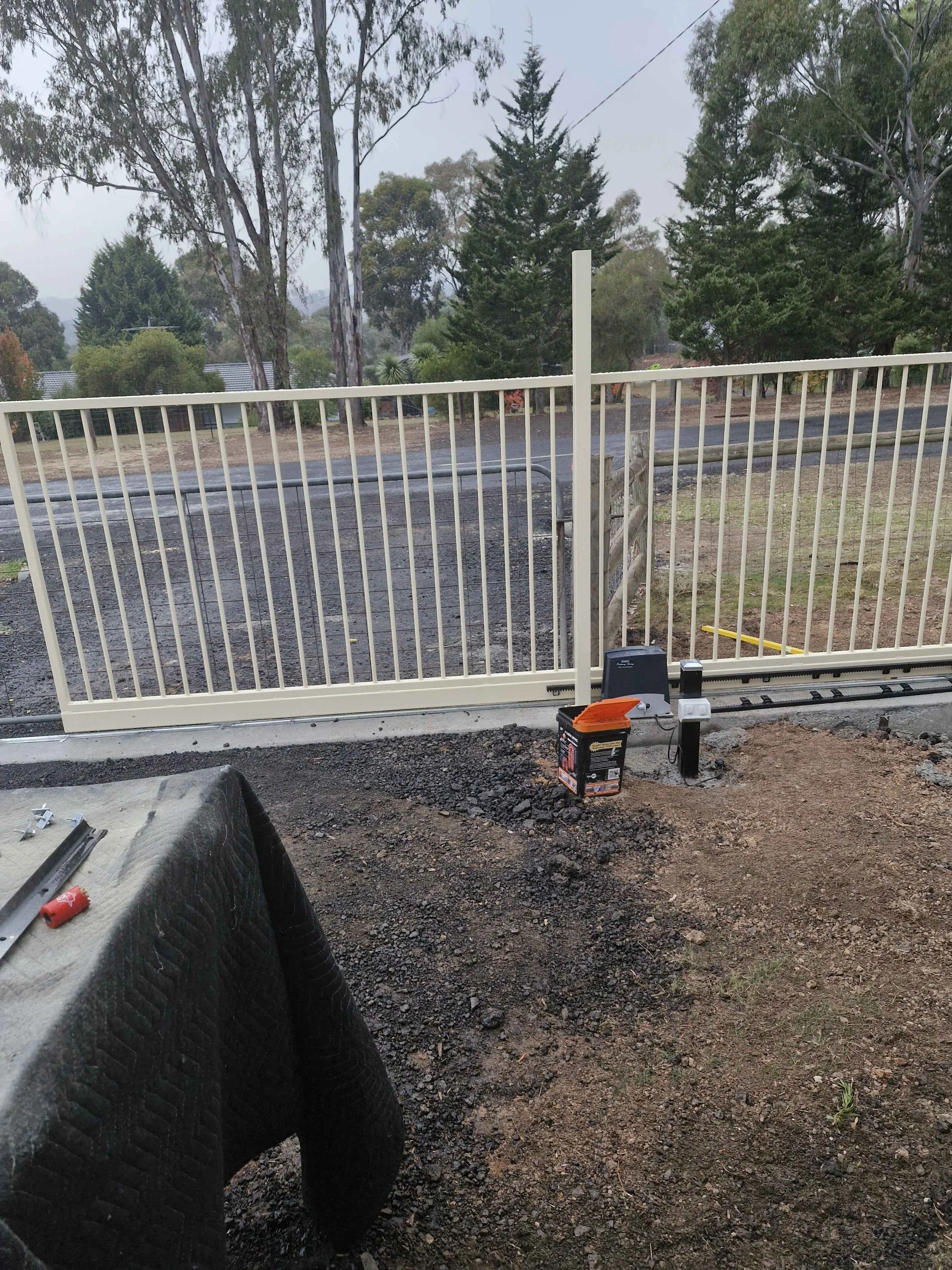 A driveway gate installation process with a partially installed white metal sliding gate, electrical equipment, and tools on the ground, with a background of trees and a road.