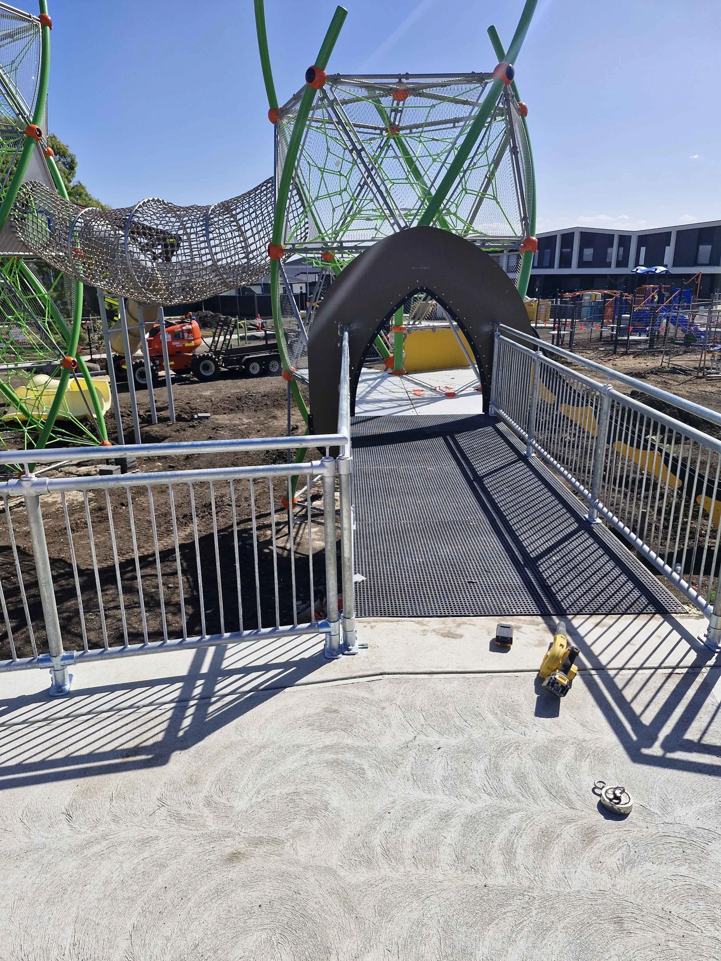 Construction site of a playground with the beginning of a metal slide and climbing structure, surrounded by metal railings, tools on the ground, and partially built equipment in the background under a clear blue sky.