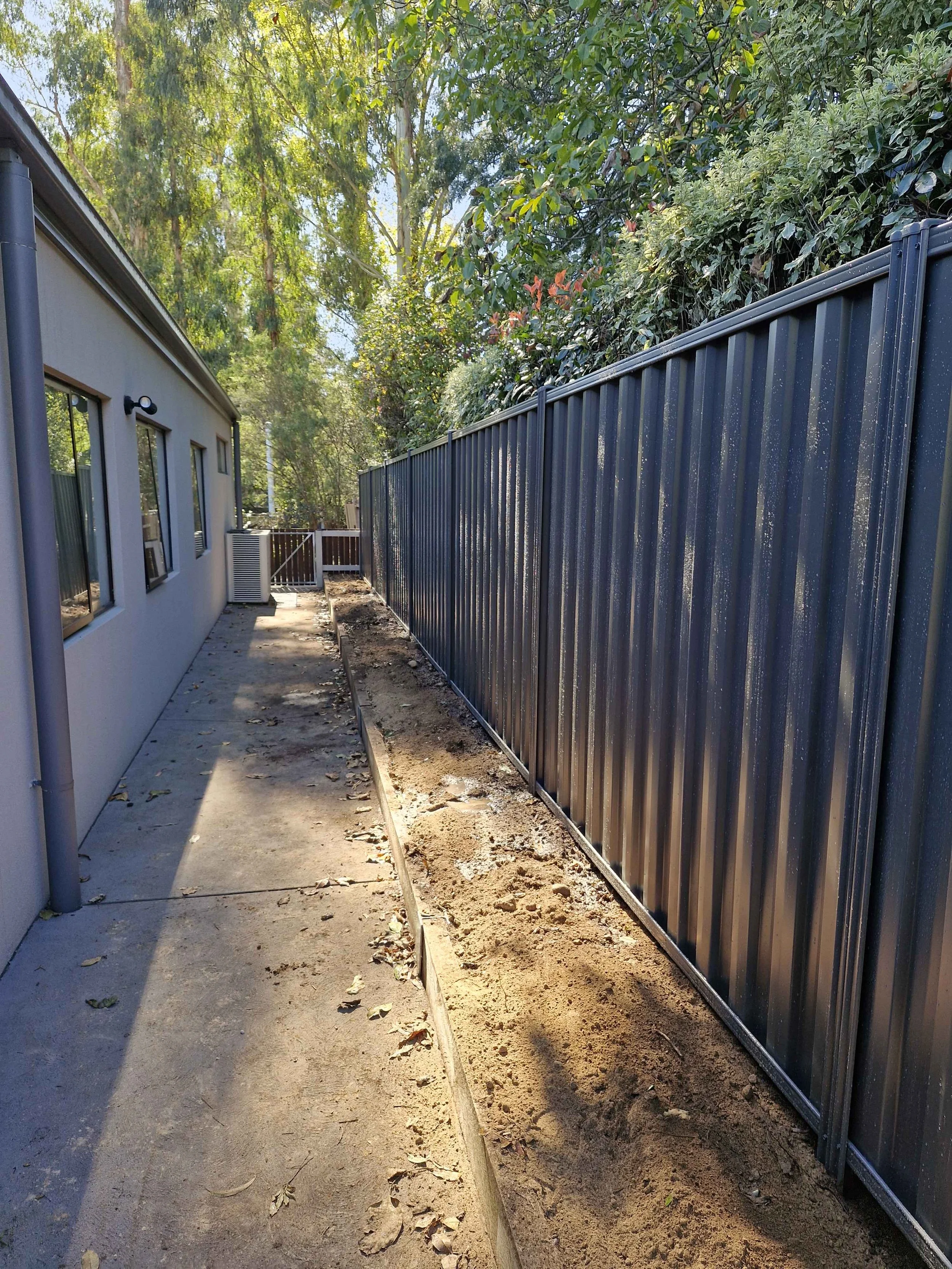 Side yard pathway with a newly installed black metal fence on the right and a house wall on the left. The pathway is partially dug up, likely for irrigation or planting, with soil and construction debris. Trees and shrubbery are visible in the backgr