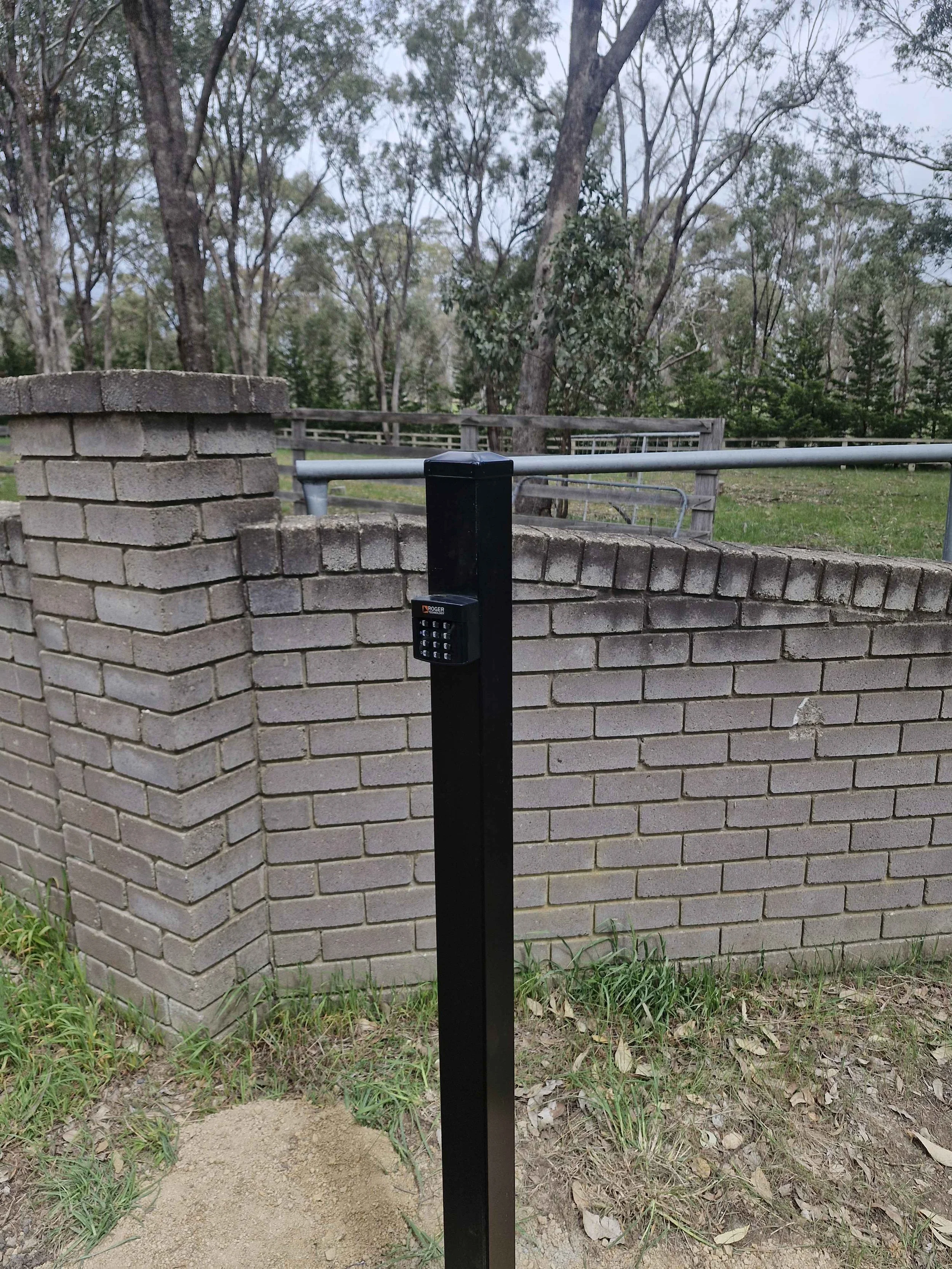 A black post with a keypad lock stands next to a brick fence with a metal gate in a park or wooded area.