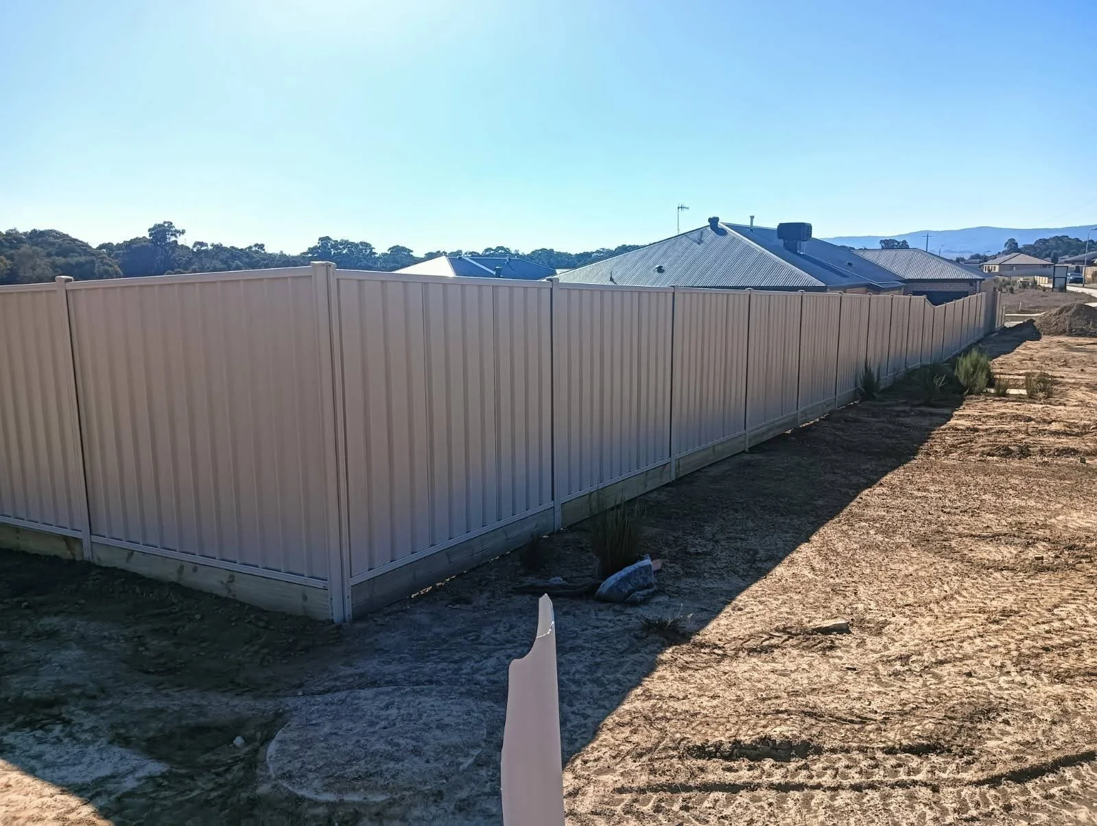 A beige metal fence encloses a backyard with dry, dusty ground, sparse plants, and a row of houses with rooftops and chimneys in the distance under clear blue skies.