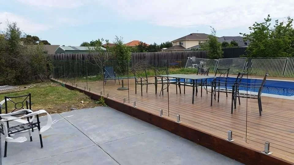 Backyard with a wooden deck around a swimming pool, patio furniture, and a glass safety fence.