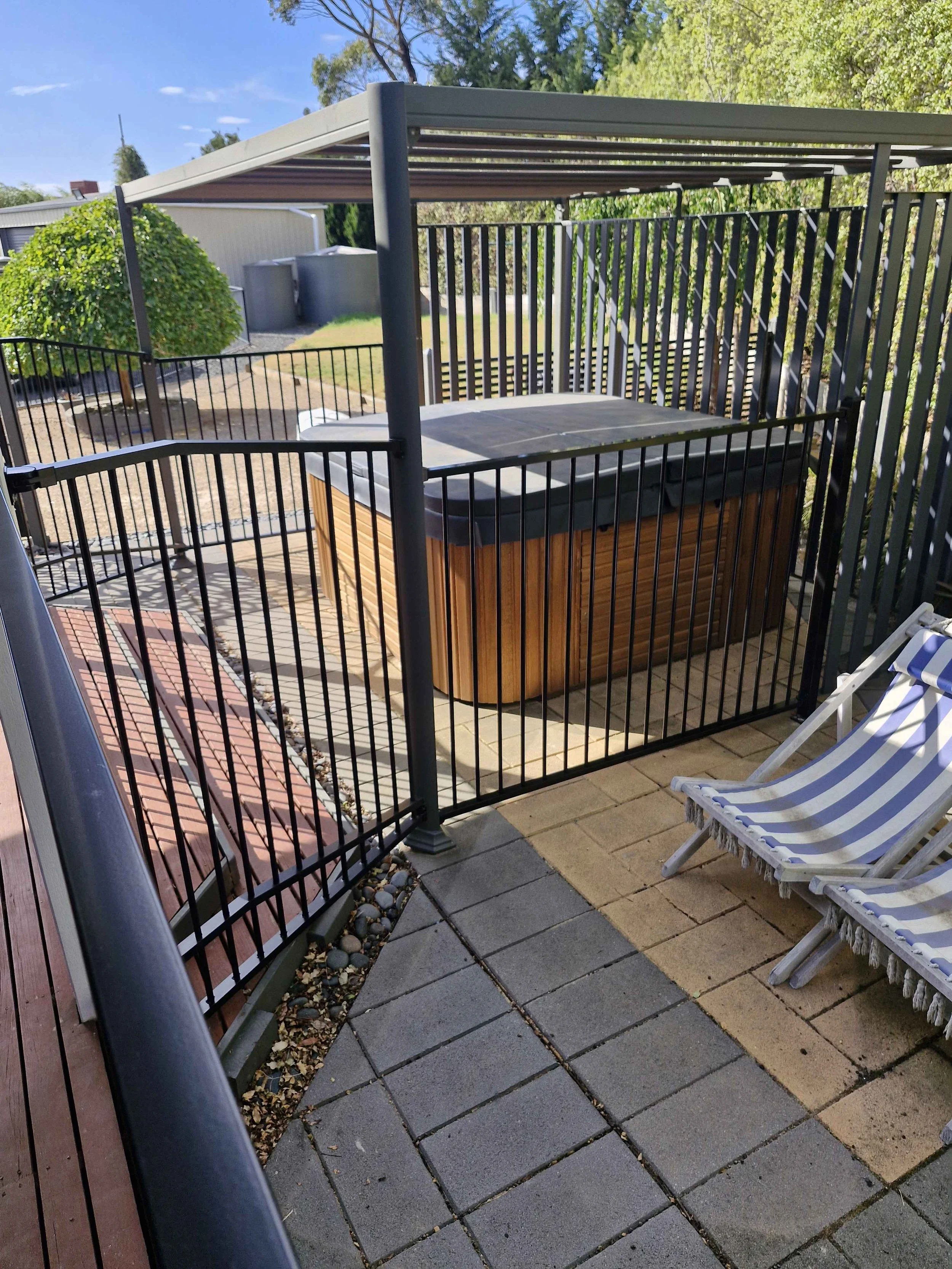 A hot tub inside a black metal fence on a patio with brick and stone pavers, adjacent to a striped fabric lounge chair with a wooden frame, and shaded by trees under a clear blue sky.