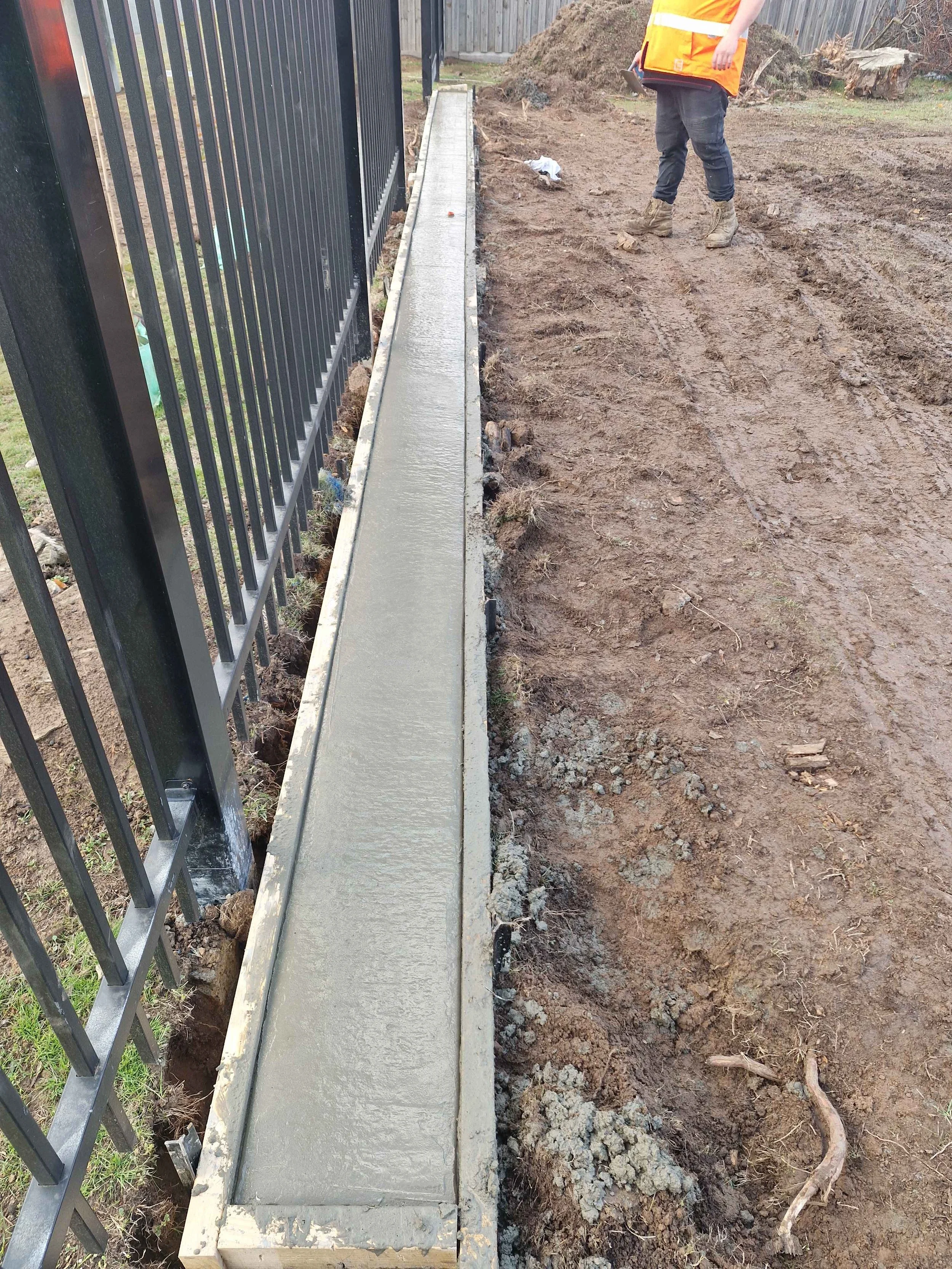 A worker wearing a safety vest and boots standing next to a freshly poured concrete sidewalk along a black metal fence on muddy ground.