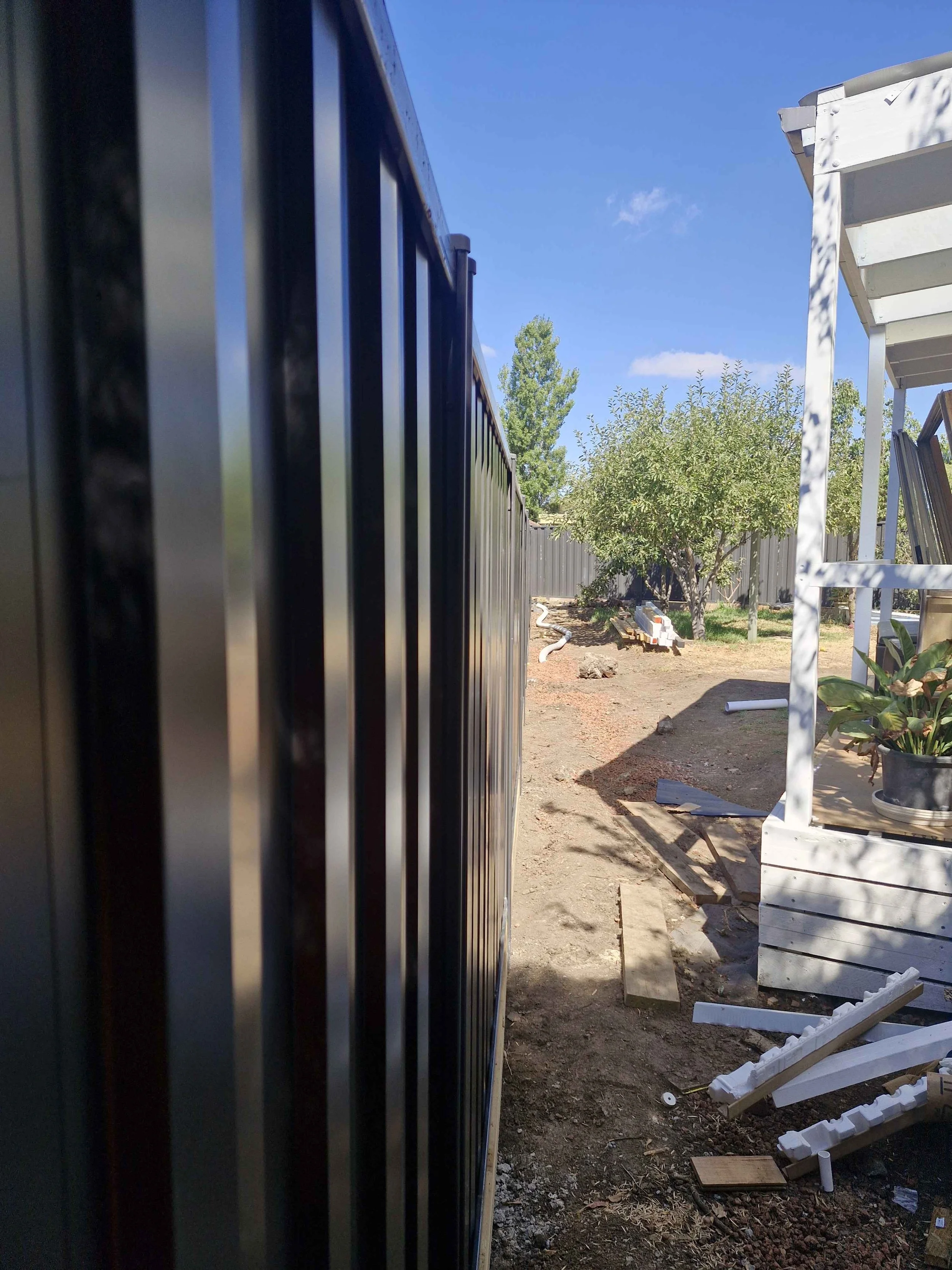 Side view of a black metal fence running along a backyard with a large tree, a white raised garden bed, and miscellaneous construction debris on the ground.