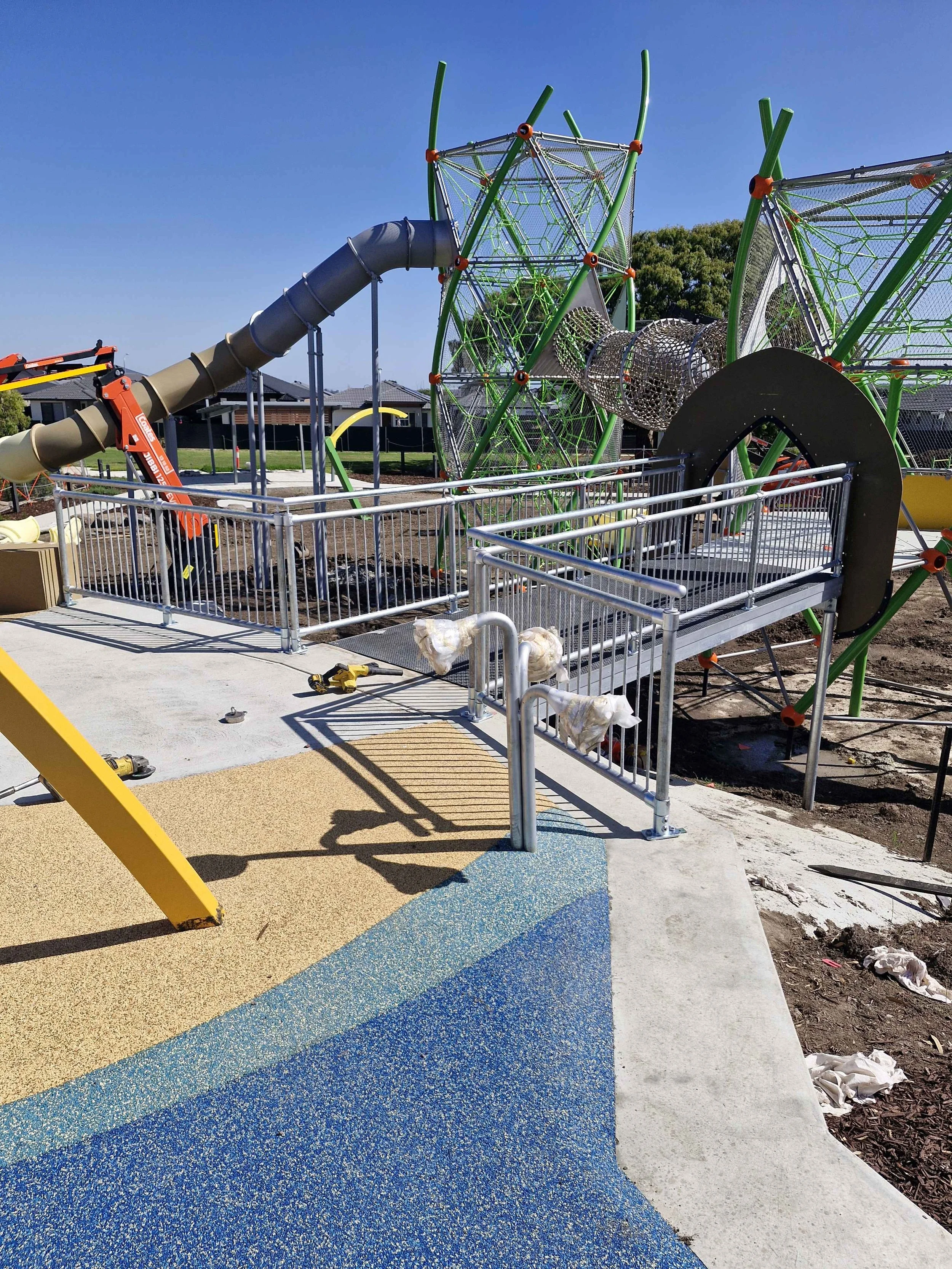 Children's playground under construction with climbing structures made of metal and plastic, slide, and surrounding safety barriers, on a bright sunny day.