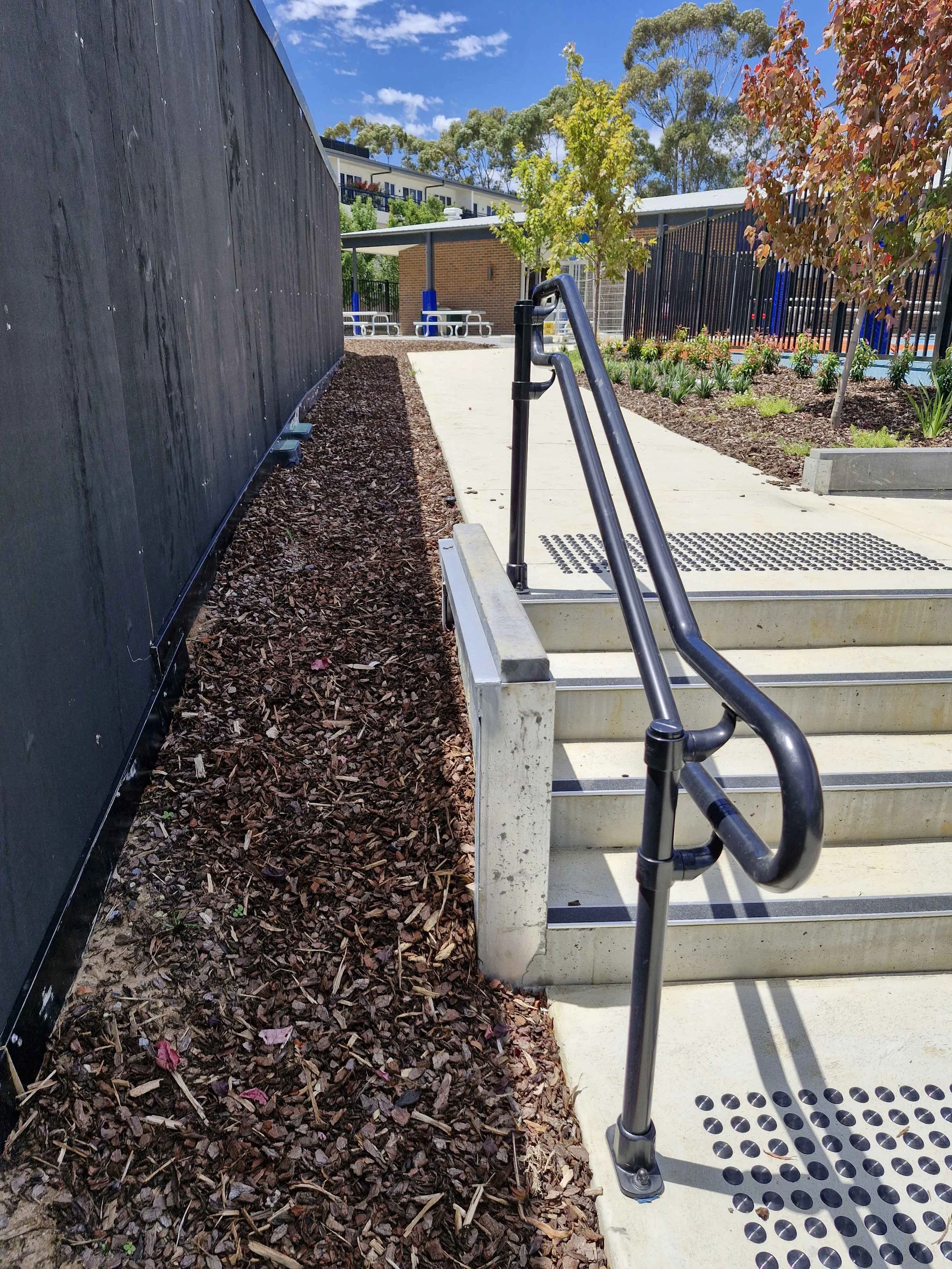 Outdoor stairway with a metal handrail leading up to a sidewalk, with mulch on the left side and landscaped planting beds with small trees and flowers on the right, in front of a black wooden fence and a building with outdoor seating.