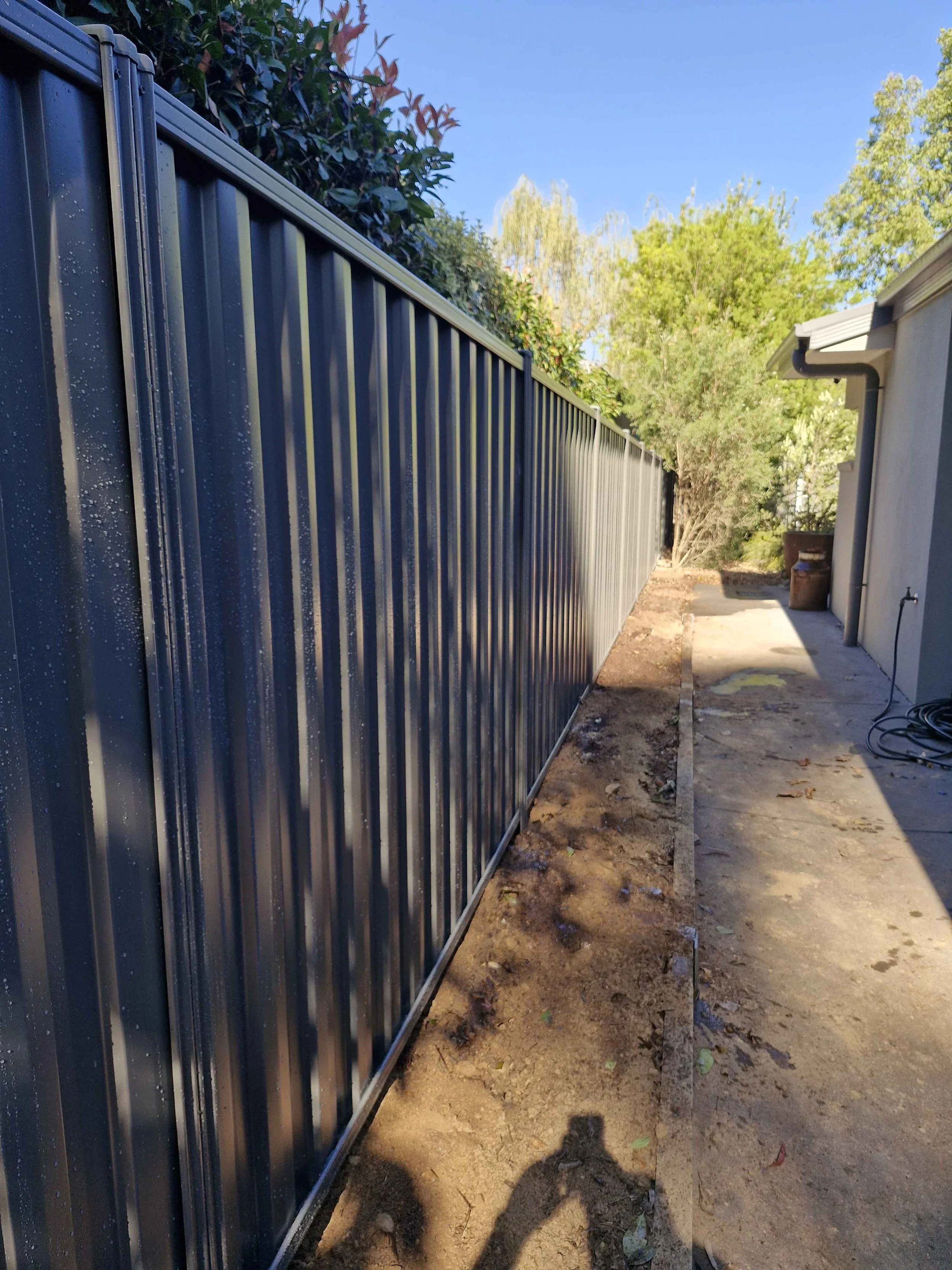 Newly installed dark metal fence along a backyard side yard with trees in the background and a house on the right.