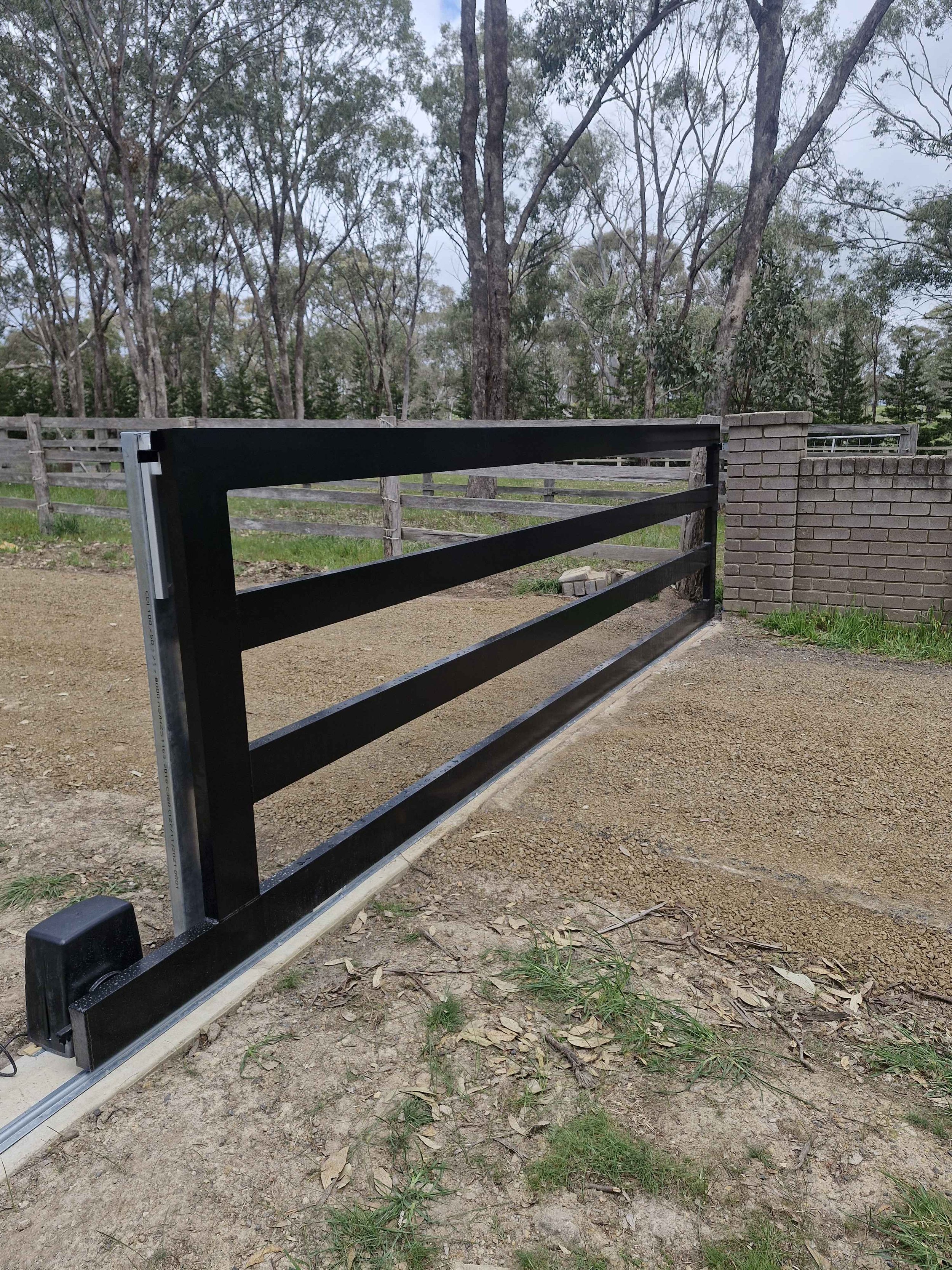 Black sliding gate installed on a dirt driveway, with a wooden fence and trees in the background.