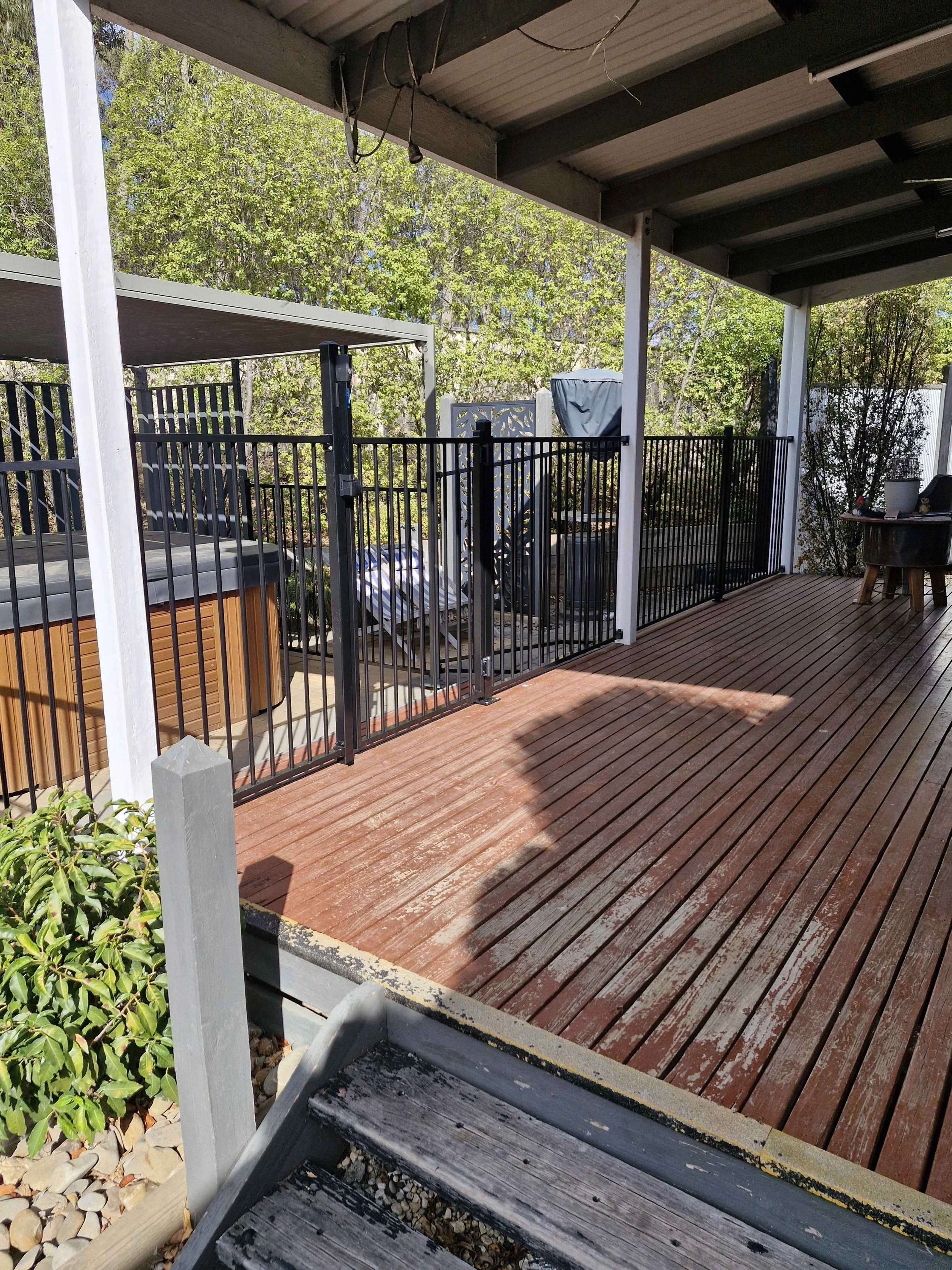 A backyard deck with wooden flooring, black metal safety gate, outdoor hot tub, and potted plants, with lush green trees in the background.