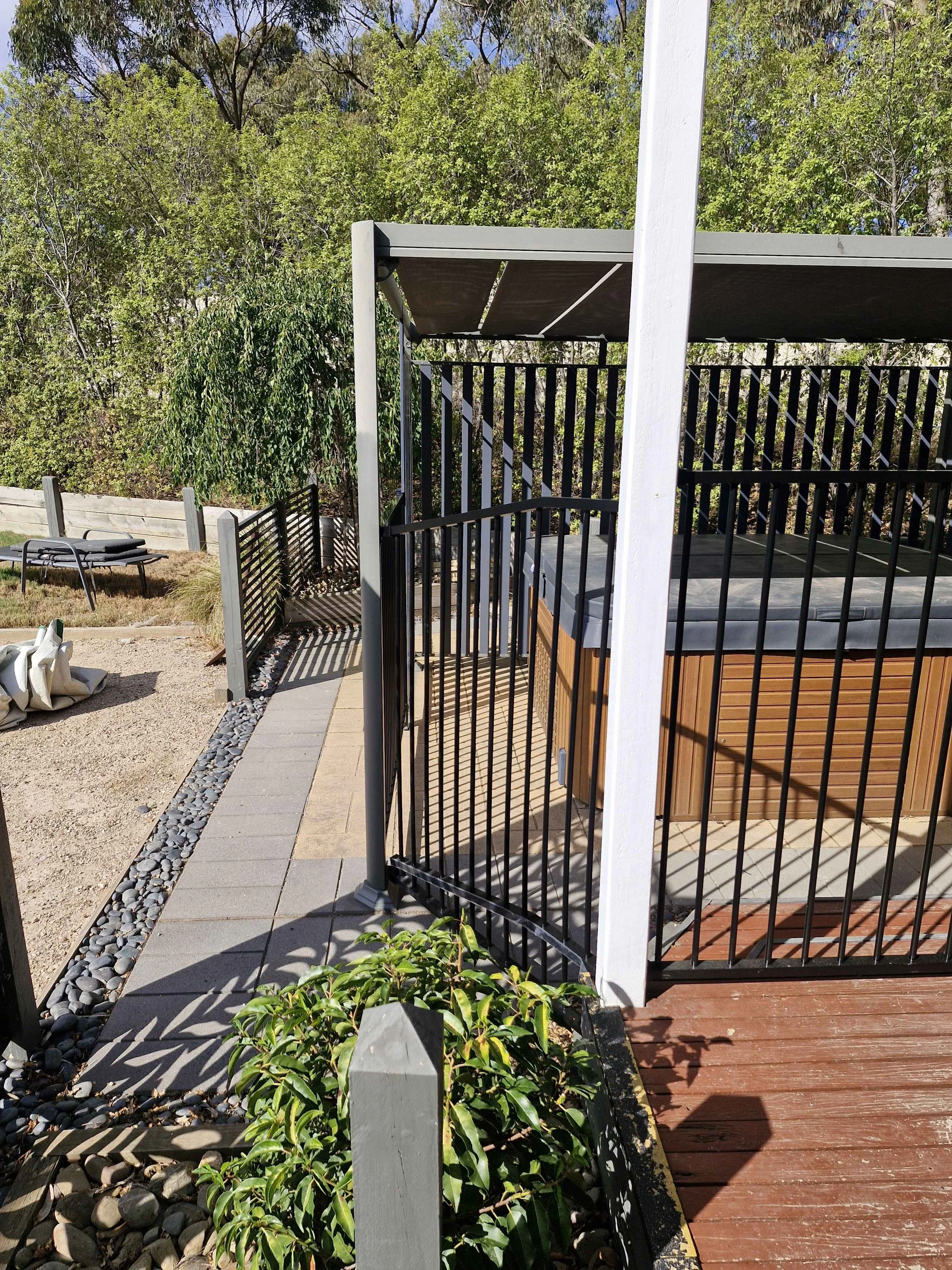Rectangular hot tub enclosed by black metal railing, situated on a wooden deck with a canopy over it. The yard has various plants, a sitting area with a lounge chair, and trees in the background.