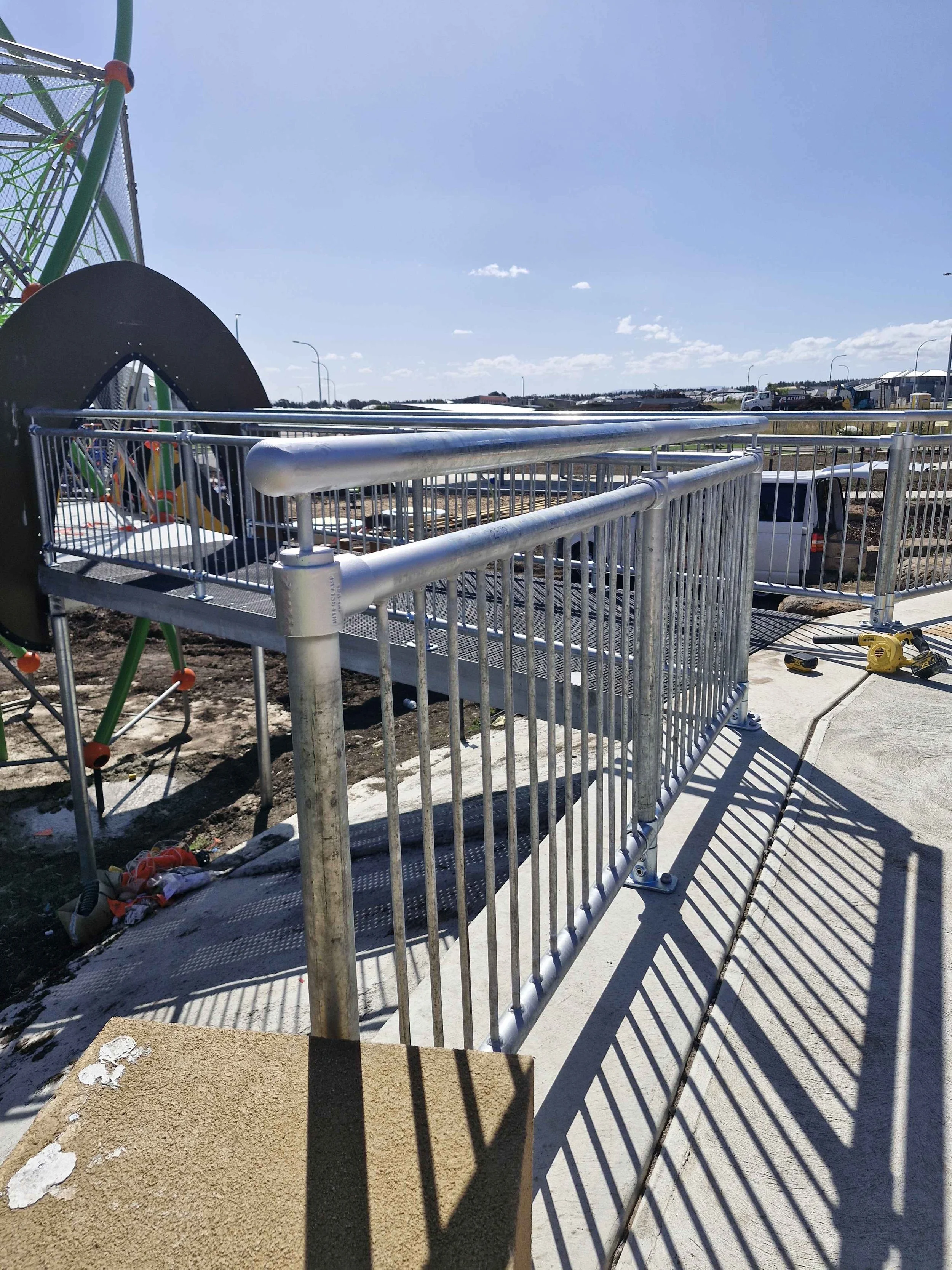 Construction site with metal railing, shadows cast on the sidewalk, playground equipment in the background, and a partly cloudy sky.