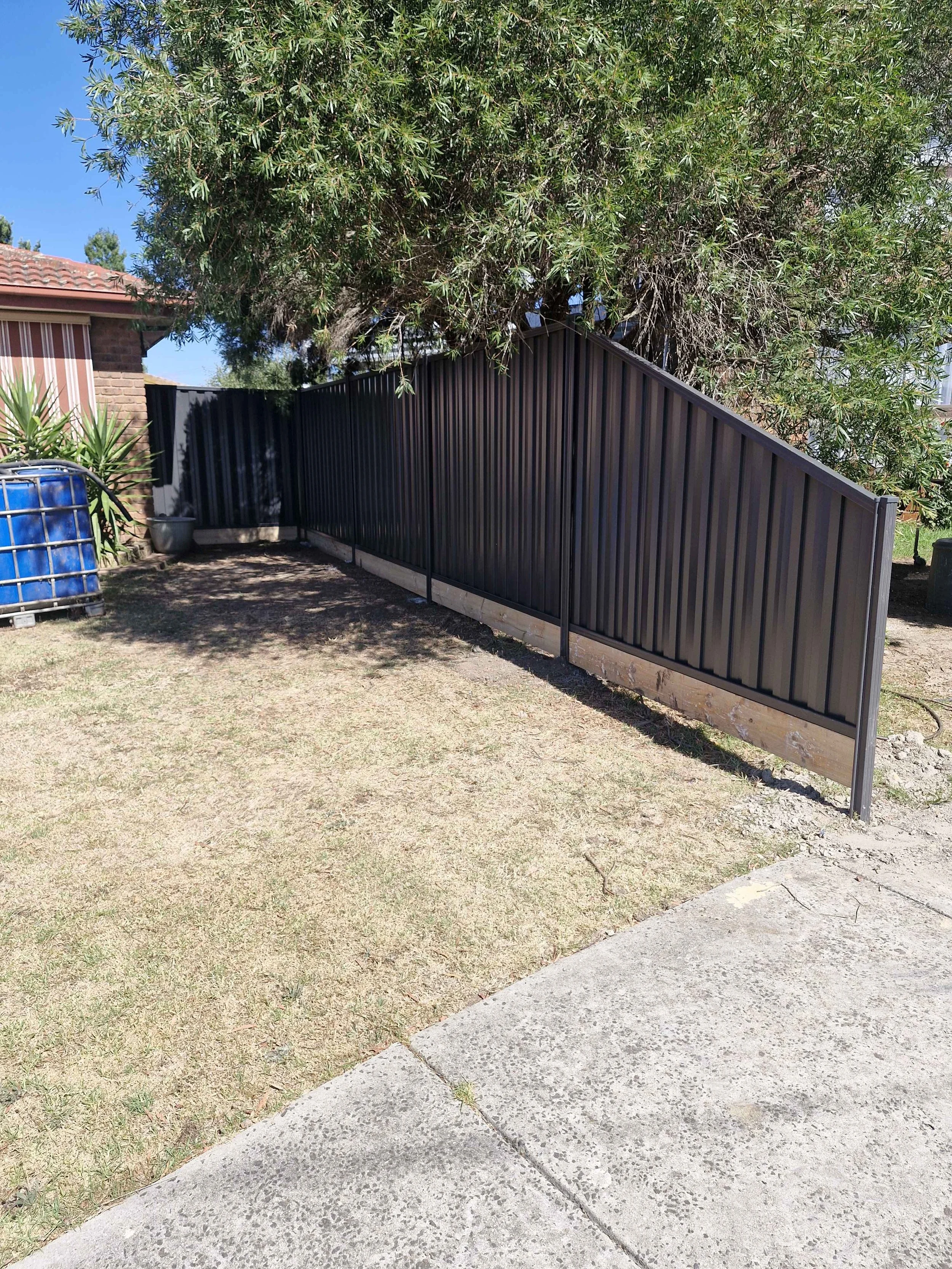 Black metal fence with wooden base in a backyard, next to a large leafy tree and a concrete pathway.