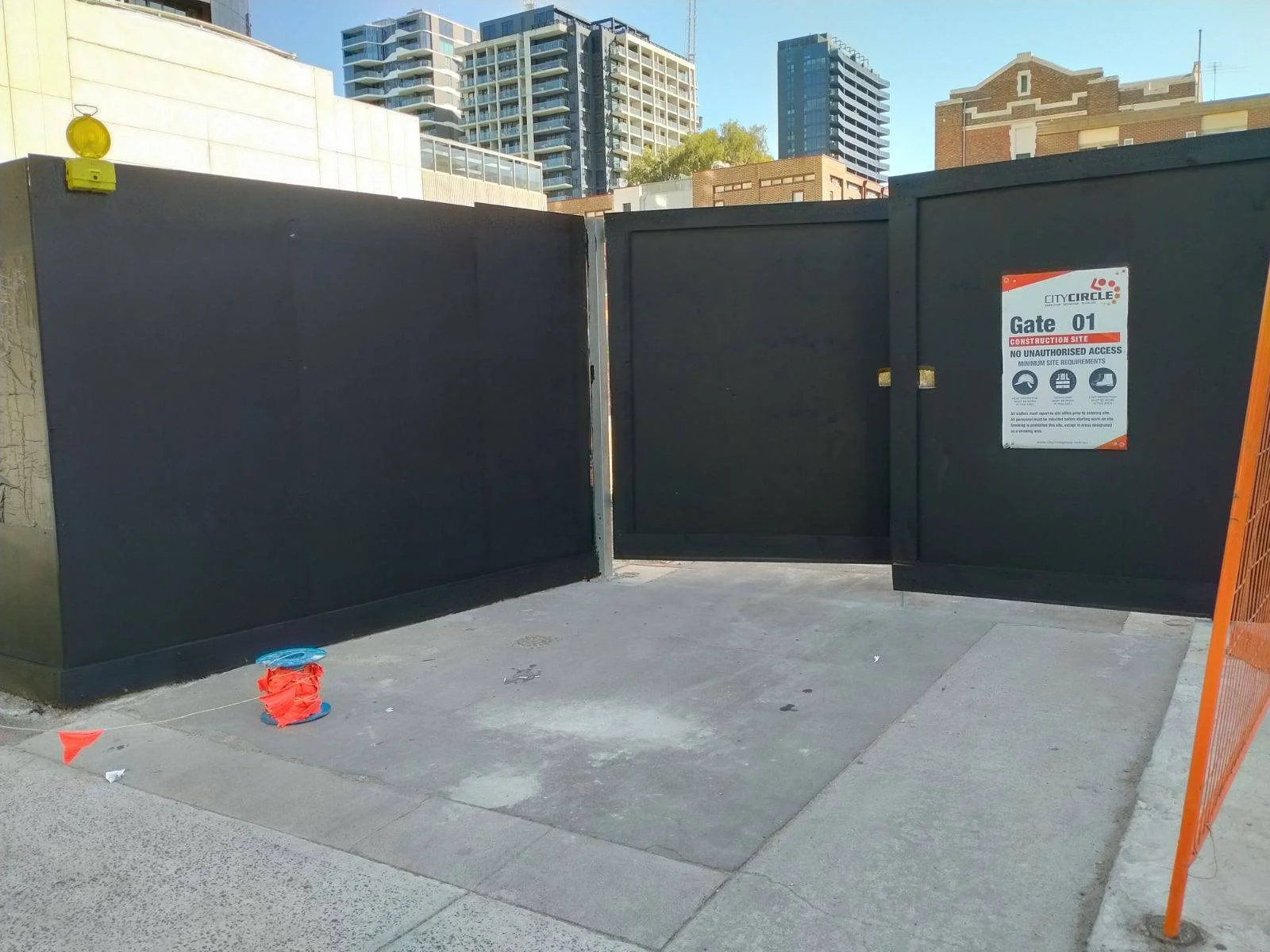 Black construction gate with a sign indicating it is Gate 01 at a construction site in an urban area, with high-rise buildings in the background.