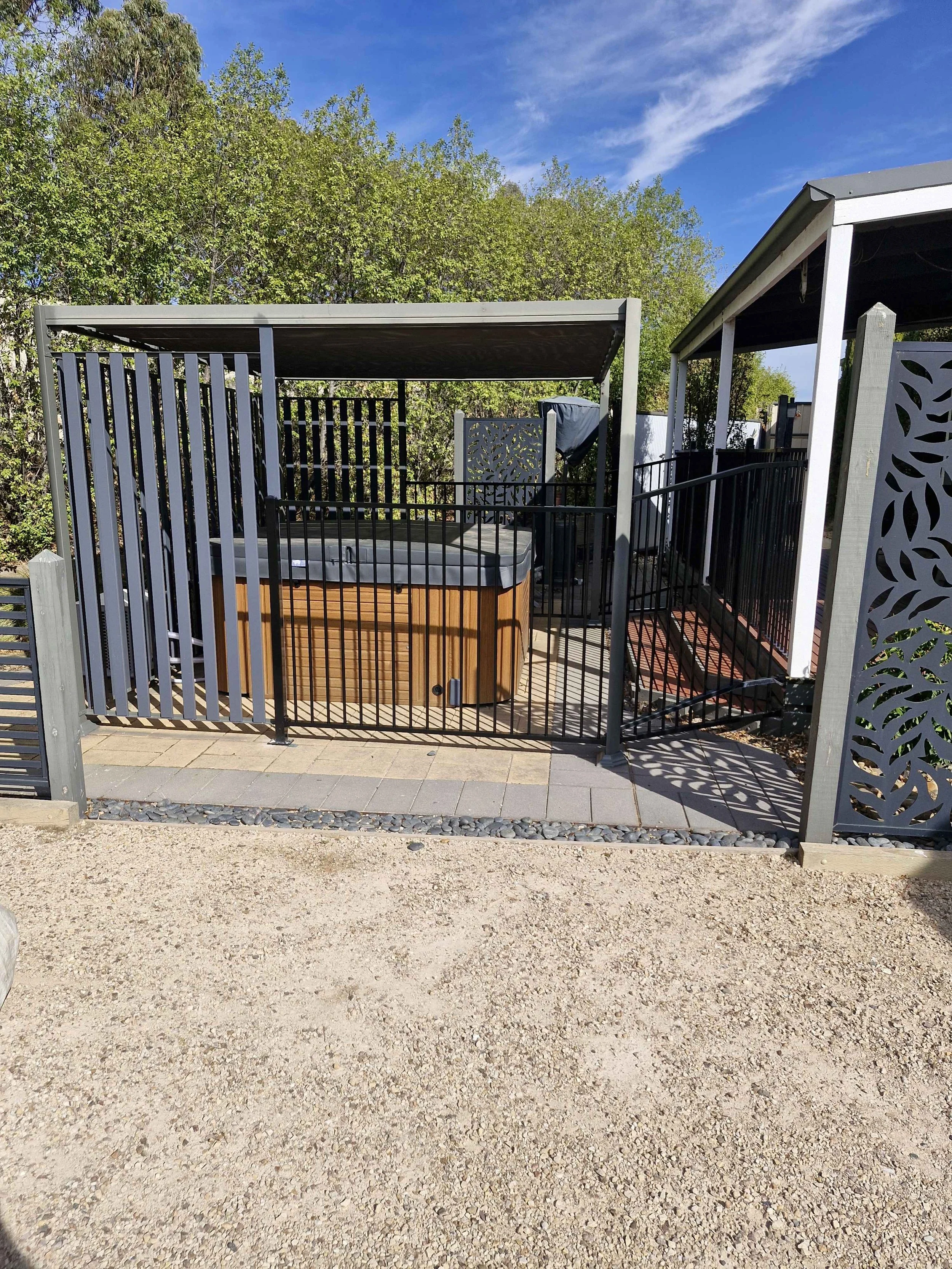 An outdoor hot tub enclosed by black and gray fencing, with a small paved area in front and a wooded background under a blue sky.