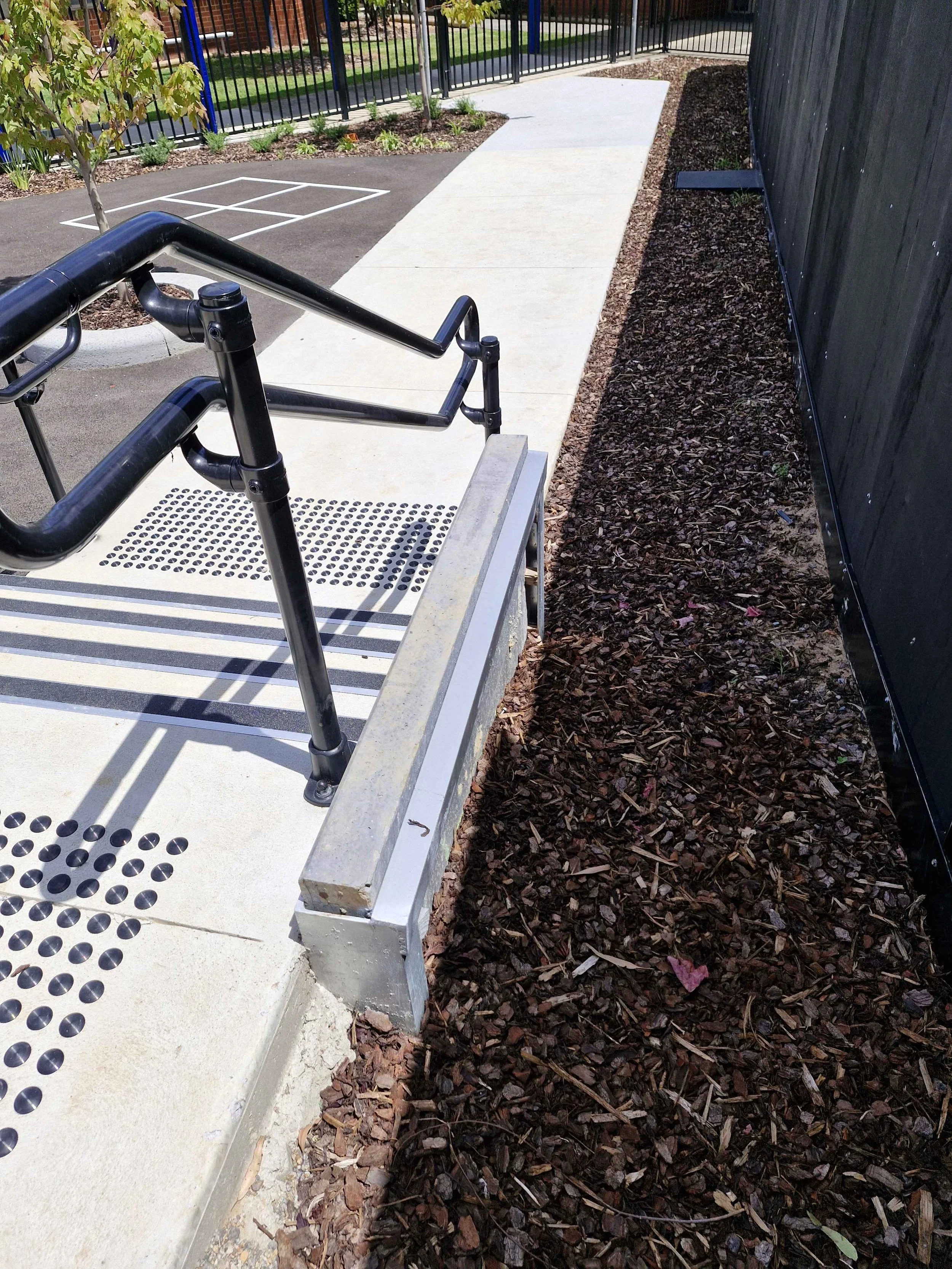 A wheelchair accessible ramp with black handrails leading to a paved walkway, bordered by mulch on the side, in a landscaped area of a park or playground.