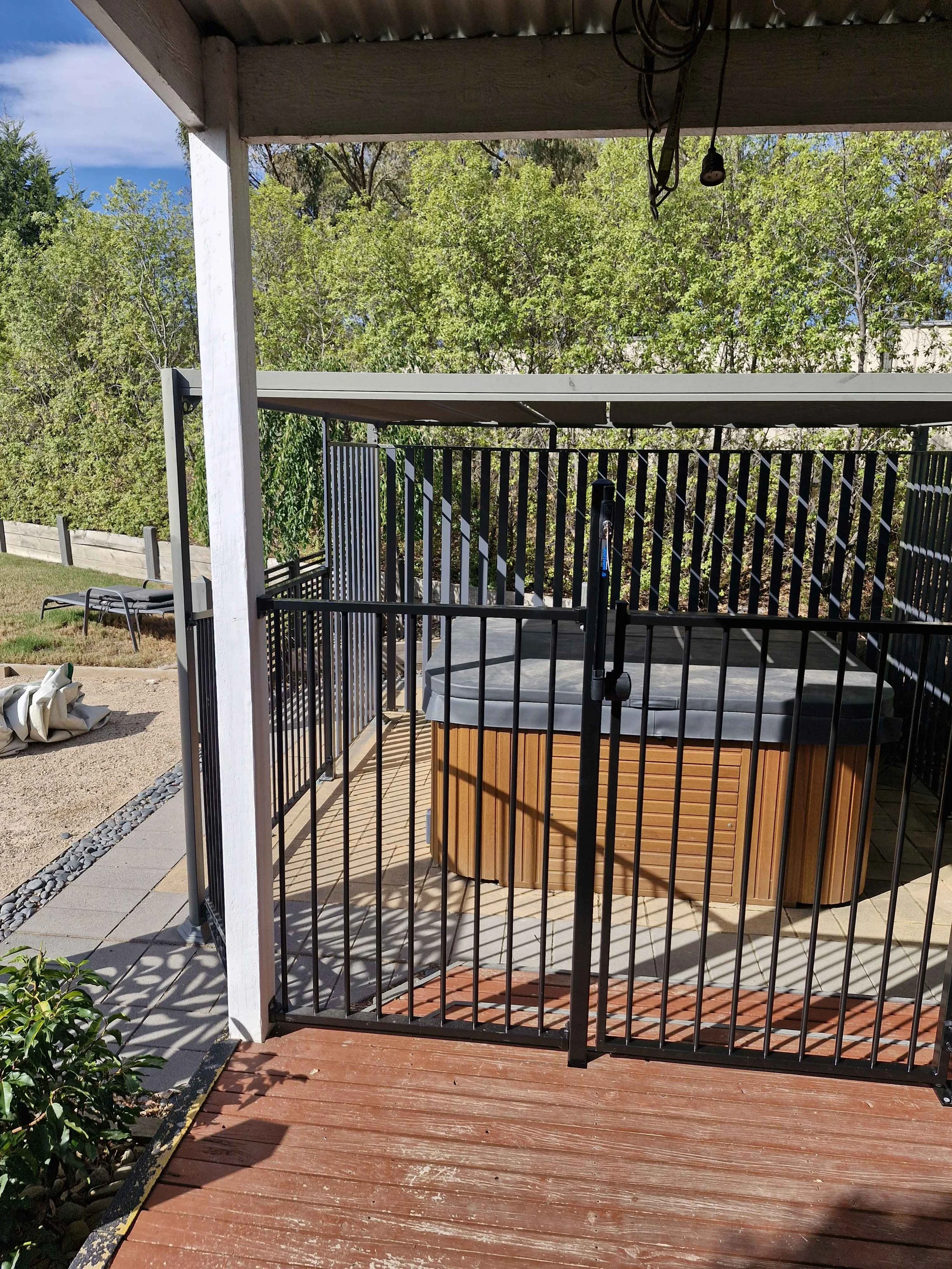 A hot tub with a black cover inside a fenced area on a wooden deck, with trees and blue sky in the background.