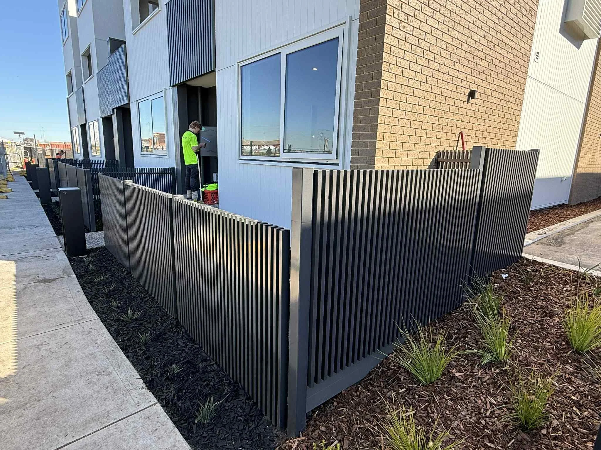 A modern apartment building with black metal fence, a sidewalk, and a man in a bright green shirt standing near a utility box.