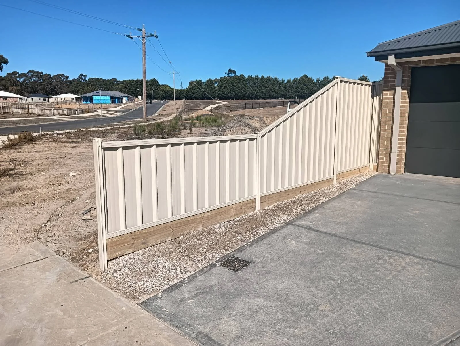 A beige fence with a wooden bottom section lines the side of a house and driveway, with a street and houses visible in the background under a clear blue sky.