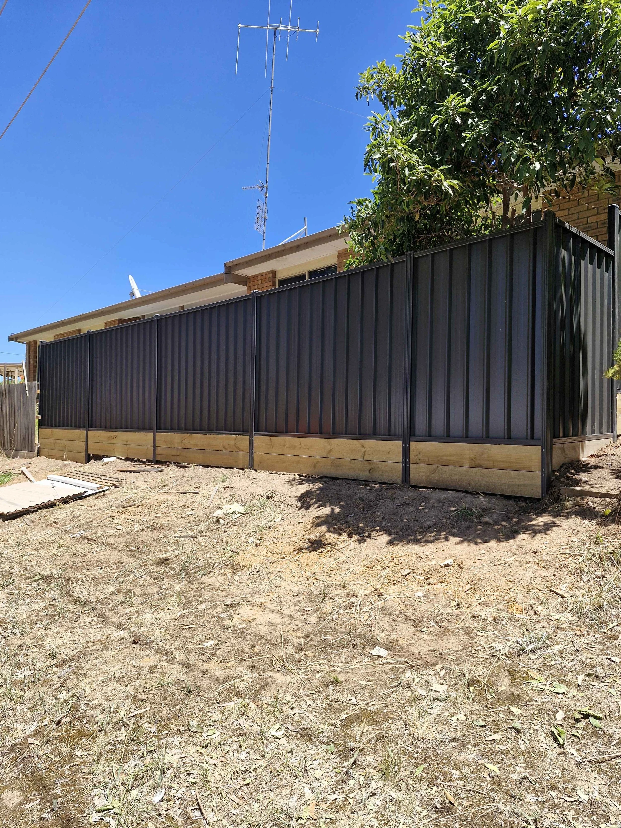 A backyard with a fixing black metal fence with wooden base, a tree, and a house with antenna on the roof, under a clear blue sky.