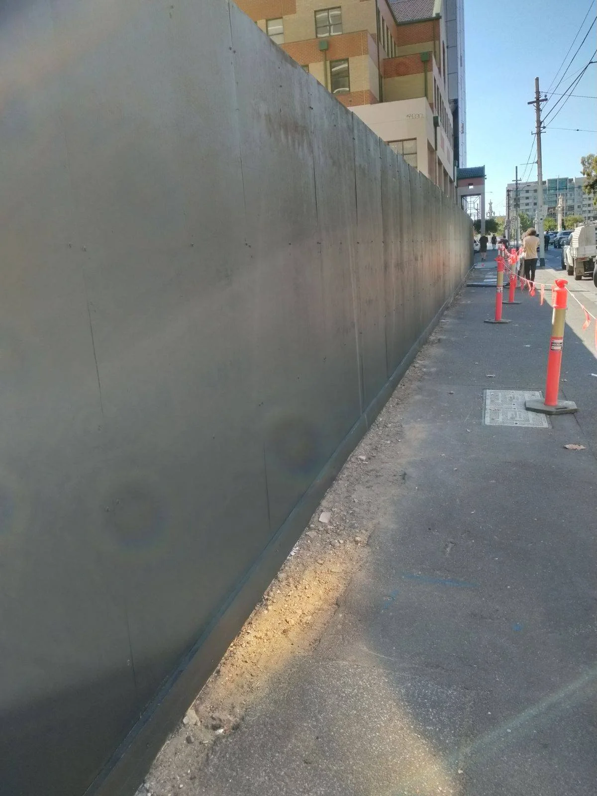 A construction site sidewalk with a tall metal fence on one side and orange traffic cones with red and white caution tape on the other side, along a city street.