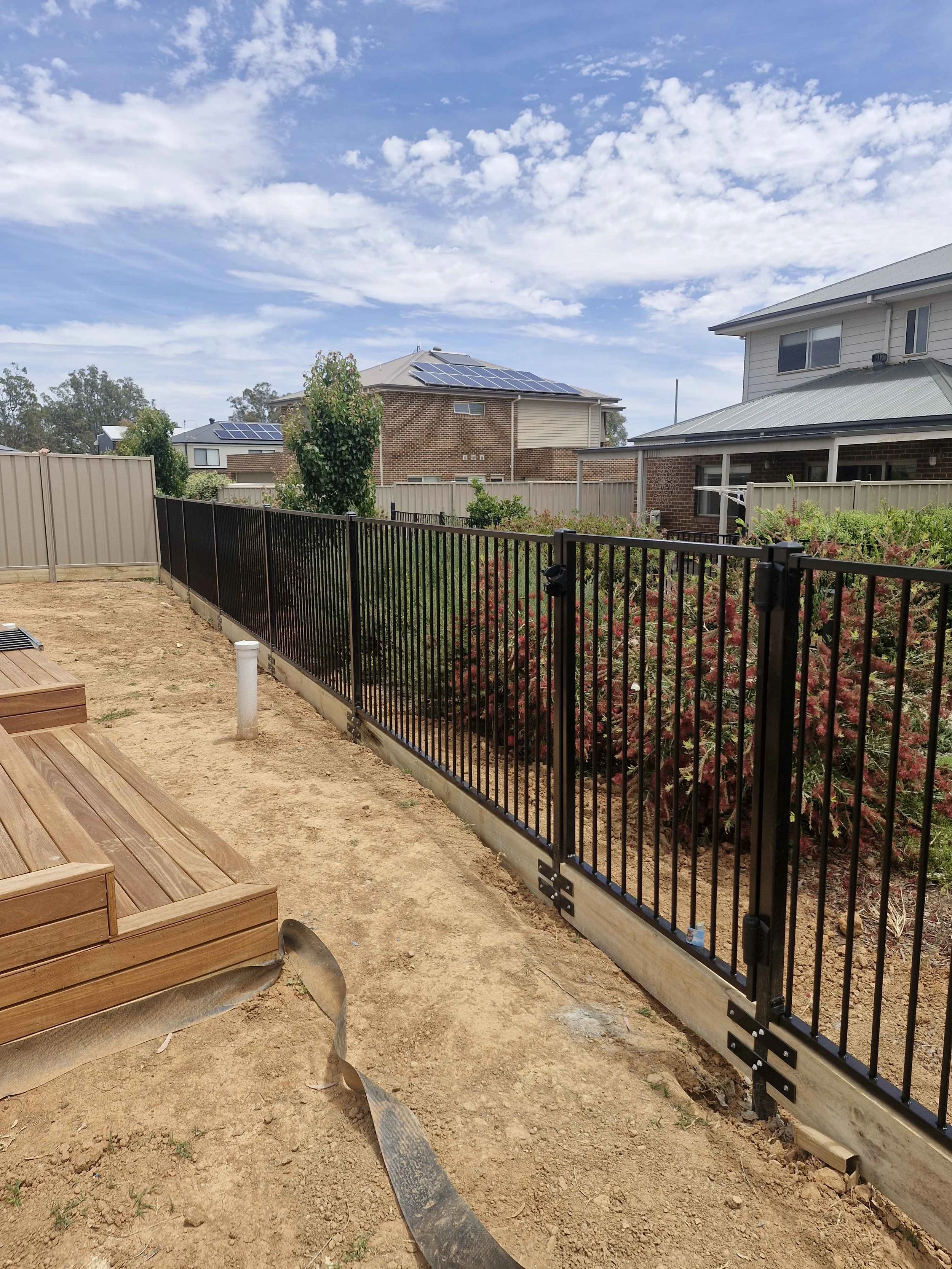 A backyard with a black metal fence, wooden deck, dirt ground, and neighboring houses with solar panels on roofs under a partly cloudy sky.