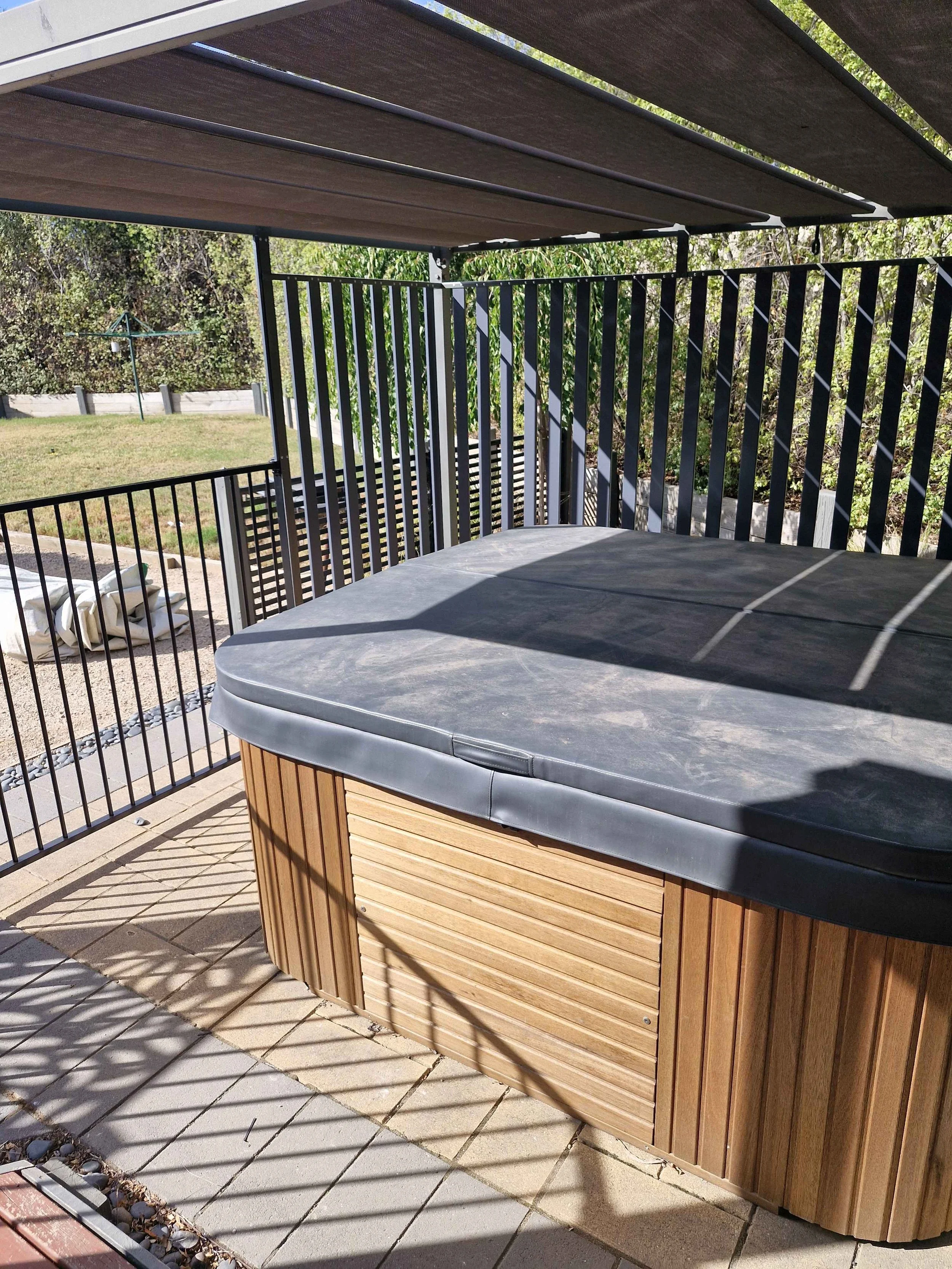 A wooden hot tub with a black cover on an outdoor patio, with a privacy fence, grassy yard, and trees in the background.