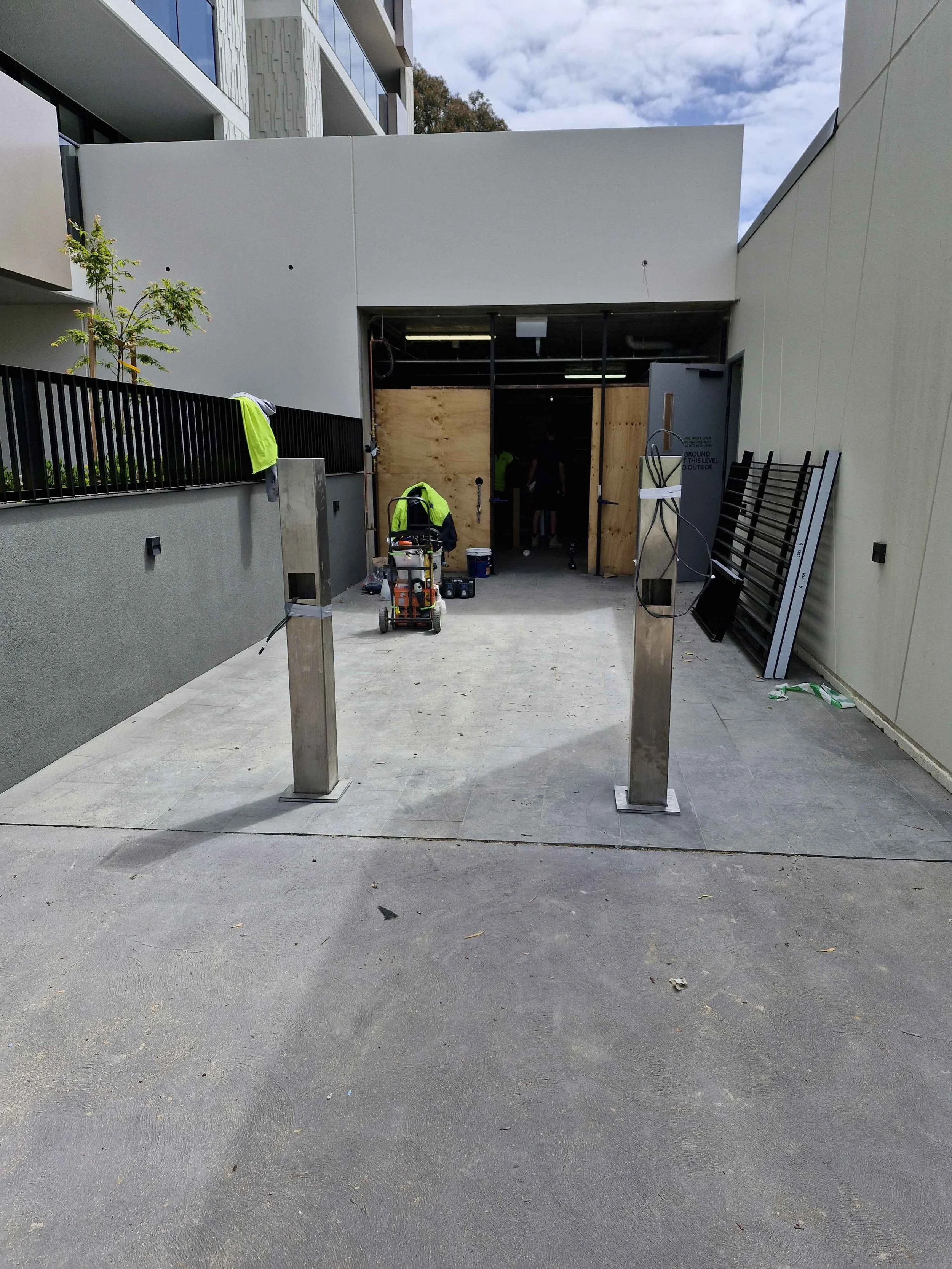 Construction site at an entrance to a building with partially installed equipment and materials, including metal posts, wooden panels, and safety vests.