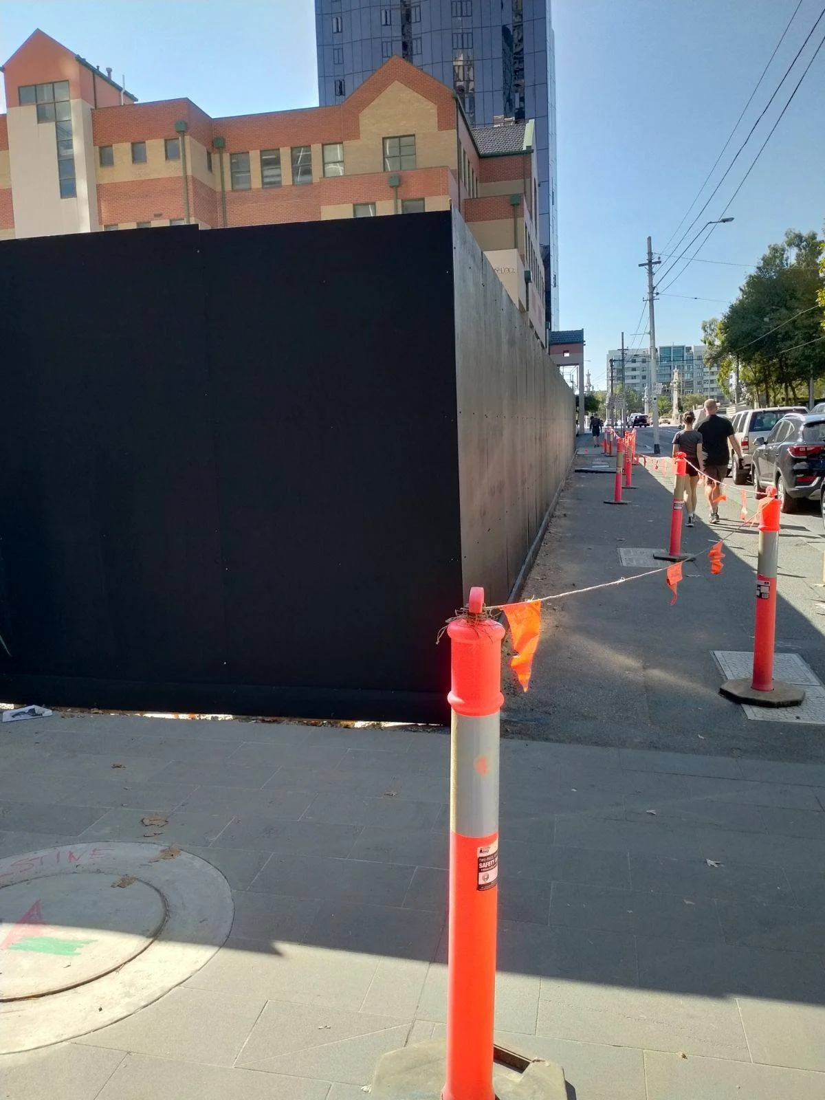 Construction area on a city sidewalk with orange safety cones and barriers, a black privacy fence, and a few pedestrians walking along the street in the background.