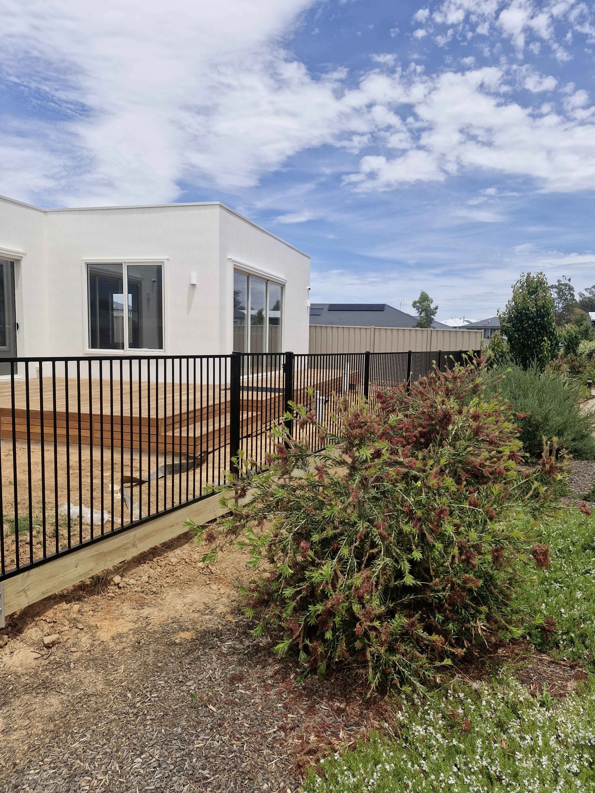 A backyard area with a modern white house, wooden deck, black metal fence, and landscaped garden plants along the fence line.