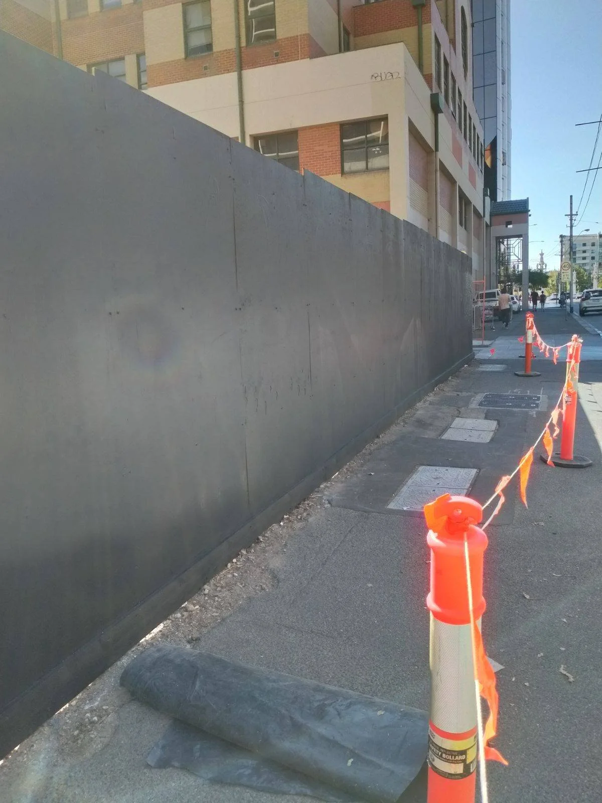 Construction site on a city sidewalk with orange safety cones and barricade tape, alongside a tall building and parked cars in the background.