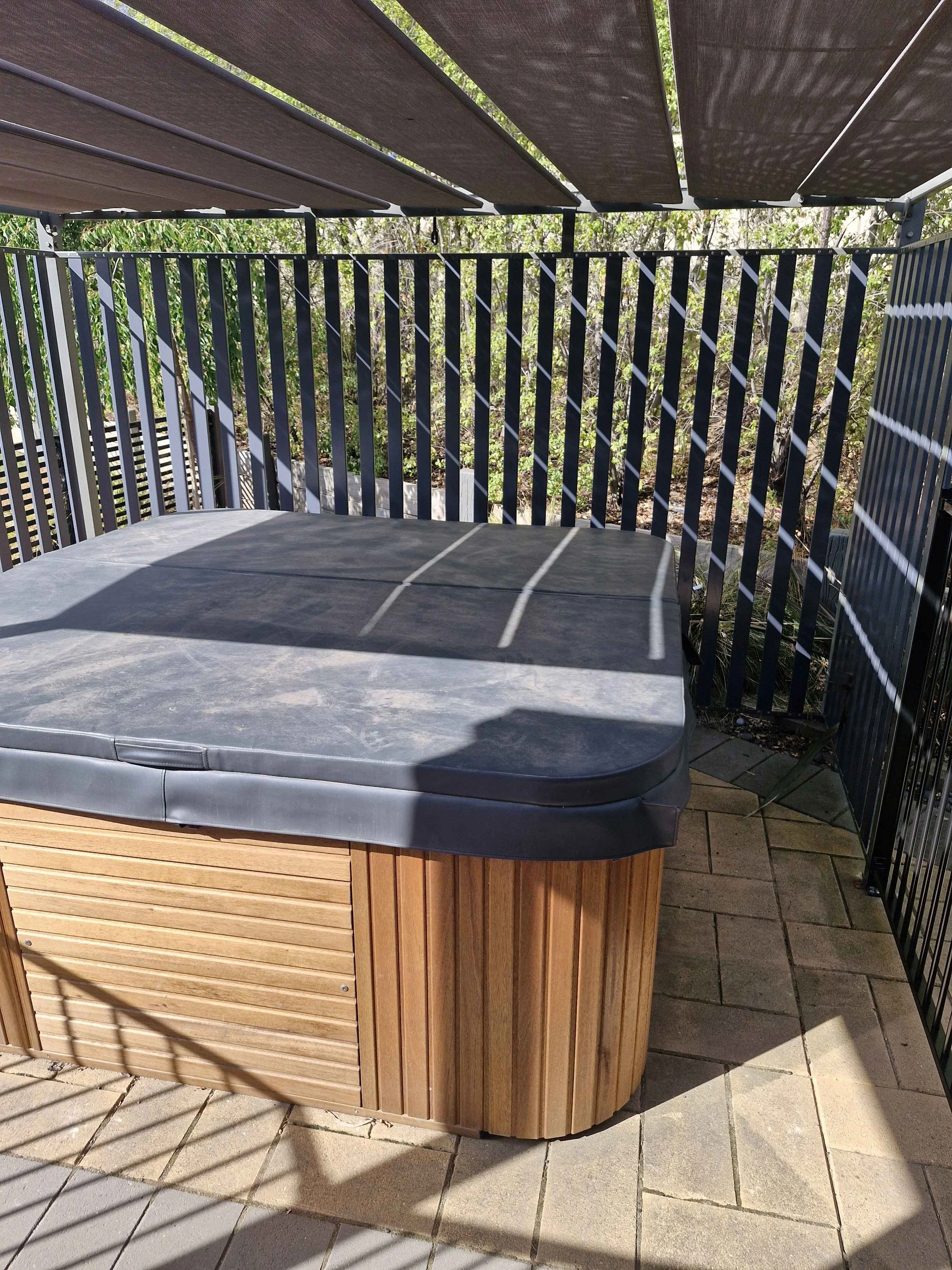 Outdoor hot tub with a gray cover on a patio, surrounded by a black metal fence and a shade sail overhead.