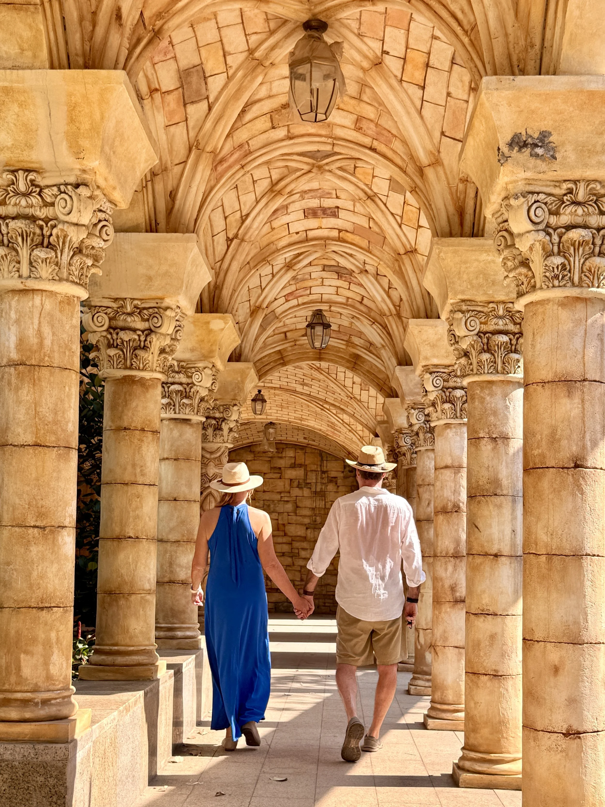 A couple holding hands while walking through a stone corridor with ornate columns and vaulted ceilings, wearing summer clothes and hats.