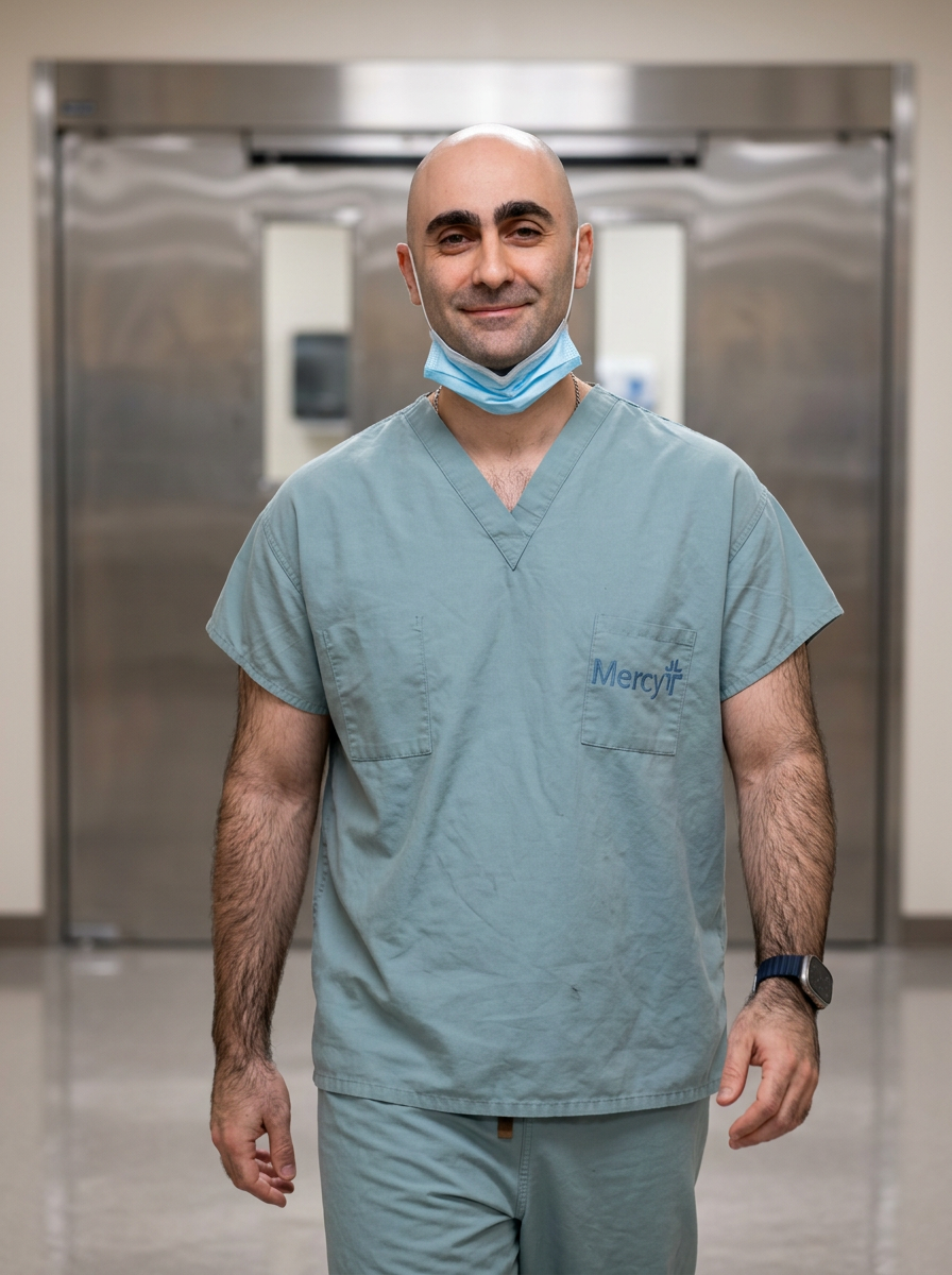 A male healthcare worker in scrubs with a blue face mask around his neck, standing in a hospital hallway with a confident smile. Surgeon physician spine spinal back neck injury hurt injured pain car accident work job pain